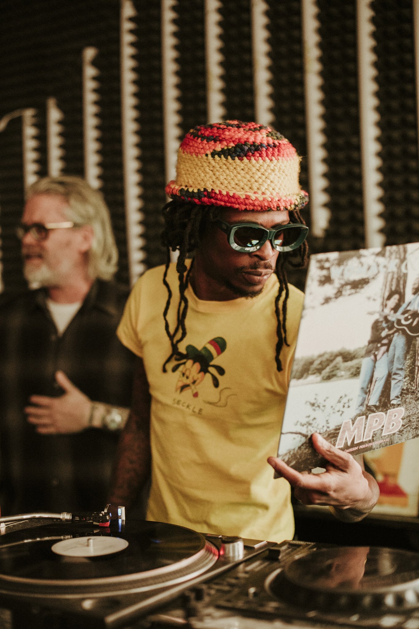 DJ holding a vinyl record in a studio, wearing a colorful knit hat and sunglasses, with another person in the background.