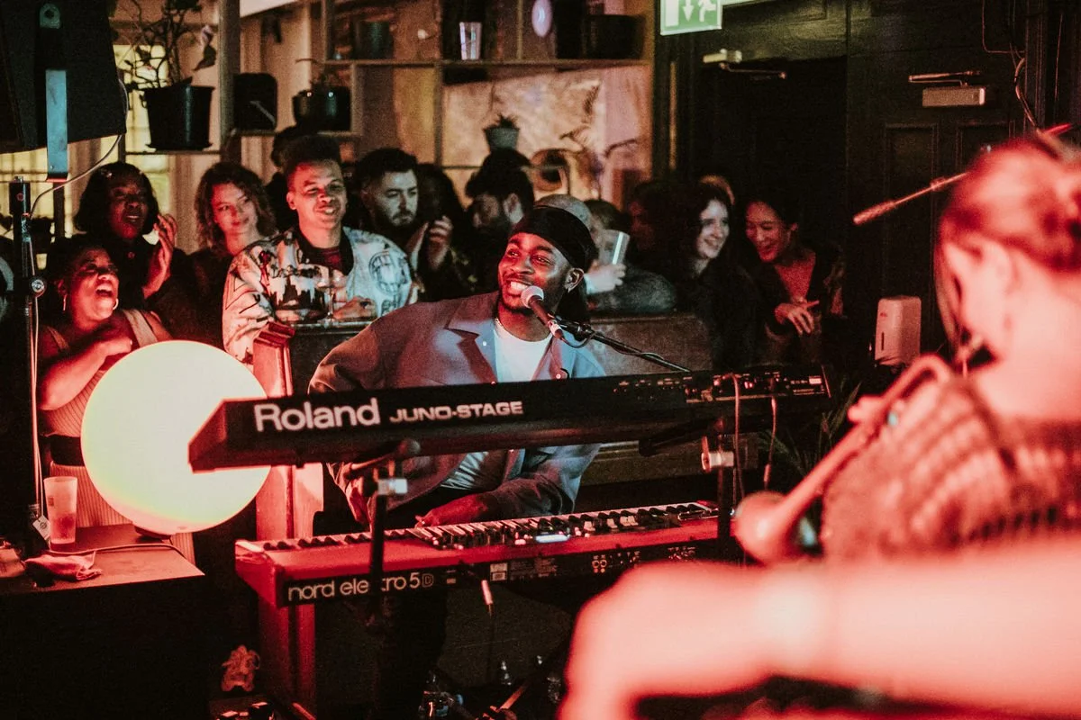 Live performance with a musician on keyboard, people watching and clapping, warm lighting.