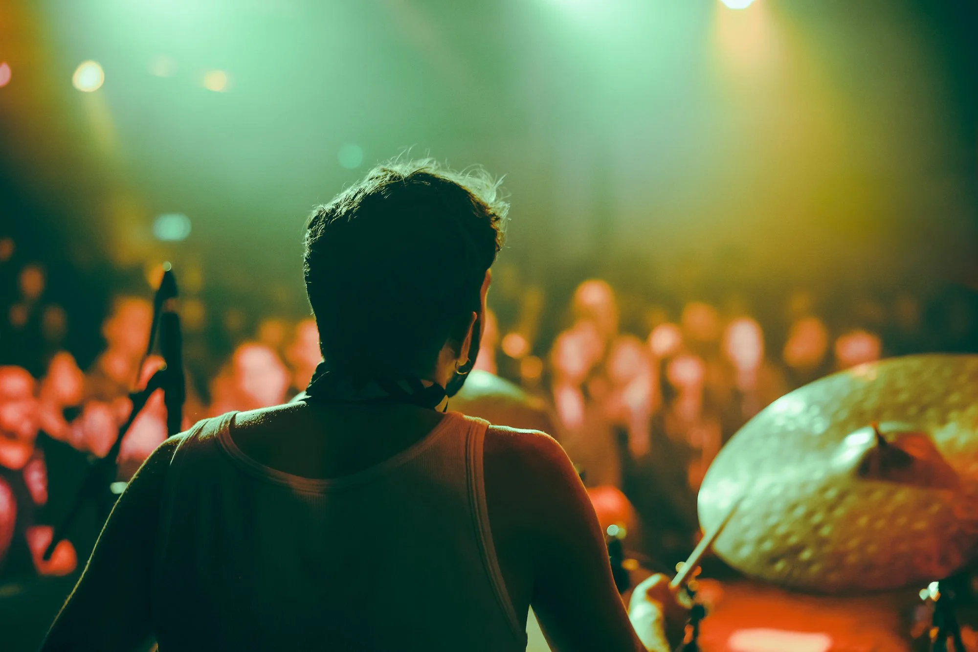 Drummer performing on stage in front of an audience with colorful concert lighting.