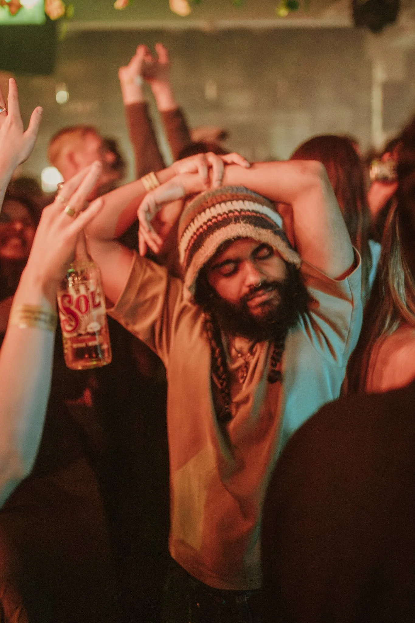 People dancing at a lively party, with colorful lighting, featuring a person in the foreground with a beanie and others holding drinks.