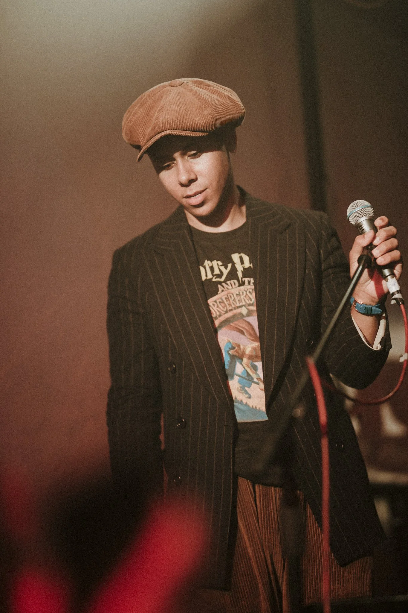 Person holding a microphone, wearing a brown cap, a pinstripe jacket, and a graphic t-shirt, standing against a brown blurred background.