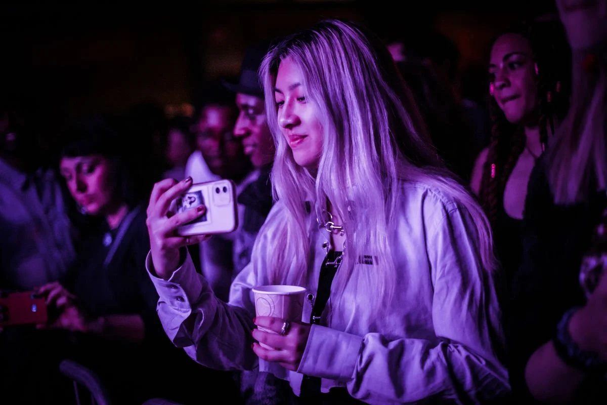 Woman with long hair taking a selfie in a dimly lit room, holding a cup, surrounded by people.