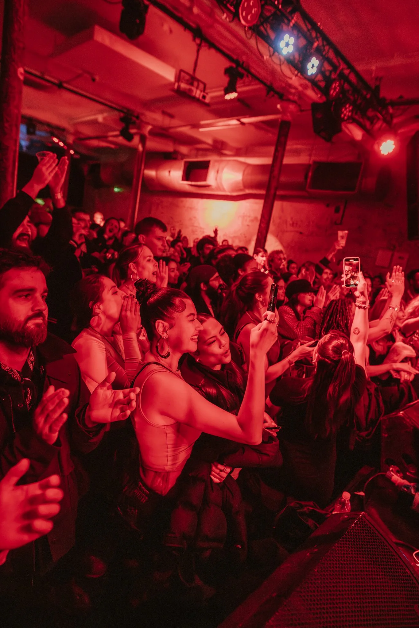 Crowd in a red-lit music venue cheering and taking photos with smartphones during a concert.