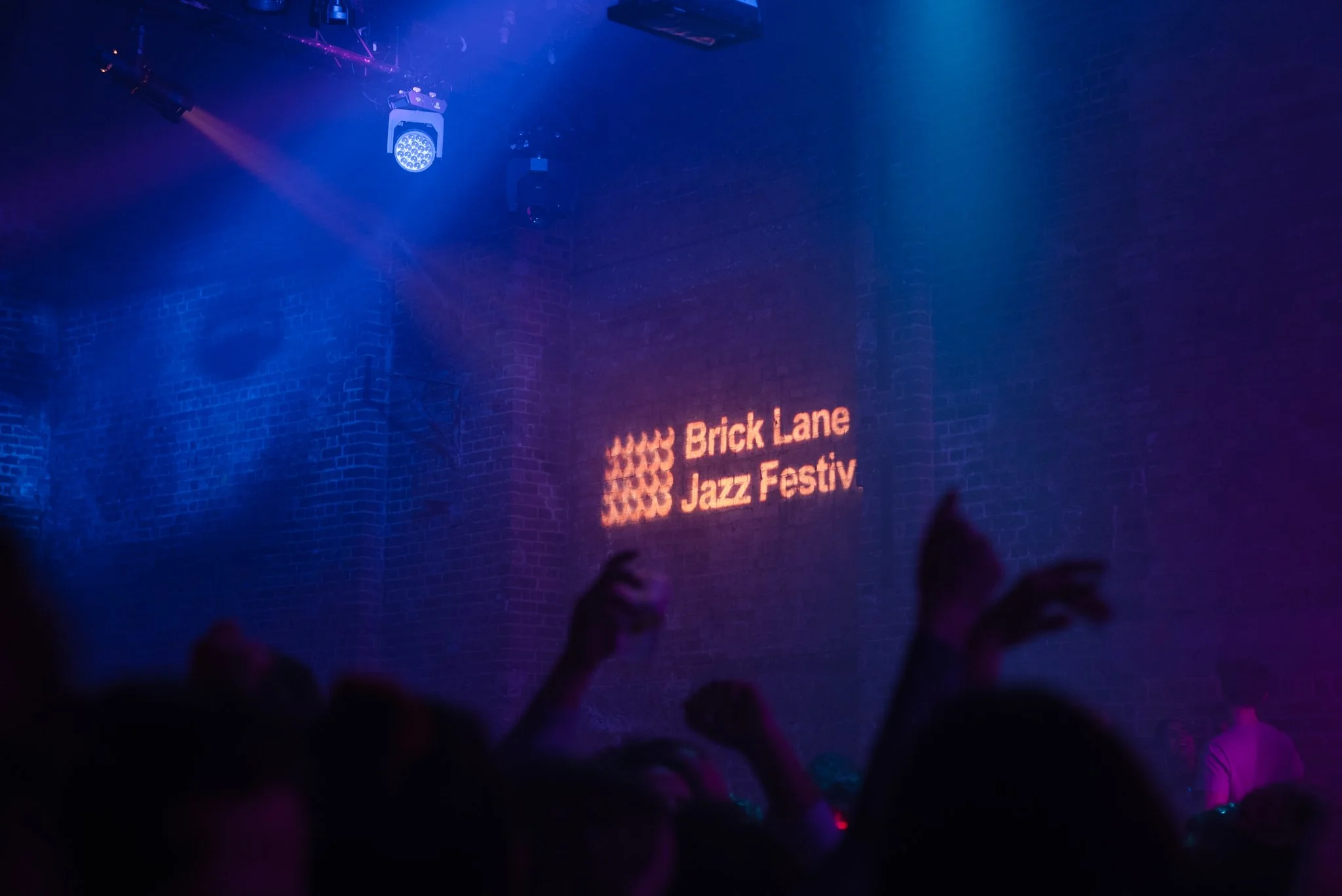 Brick Lane Jazz Festival logo projected on a wall in a dimly lit venue with silhouetted crowd.