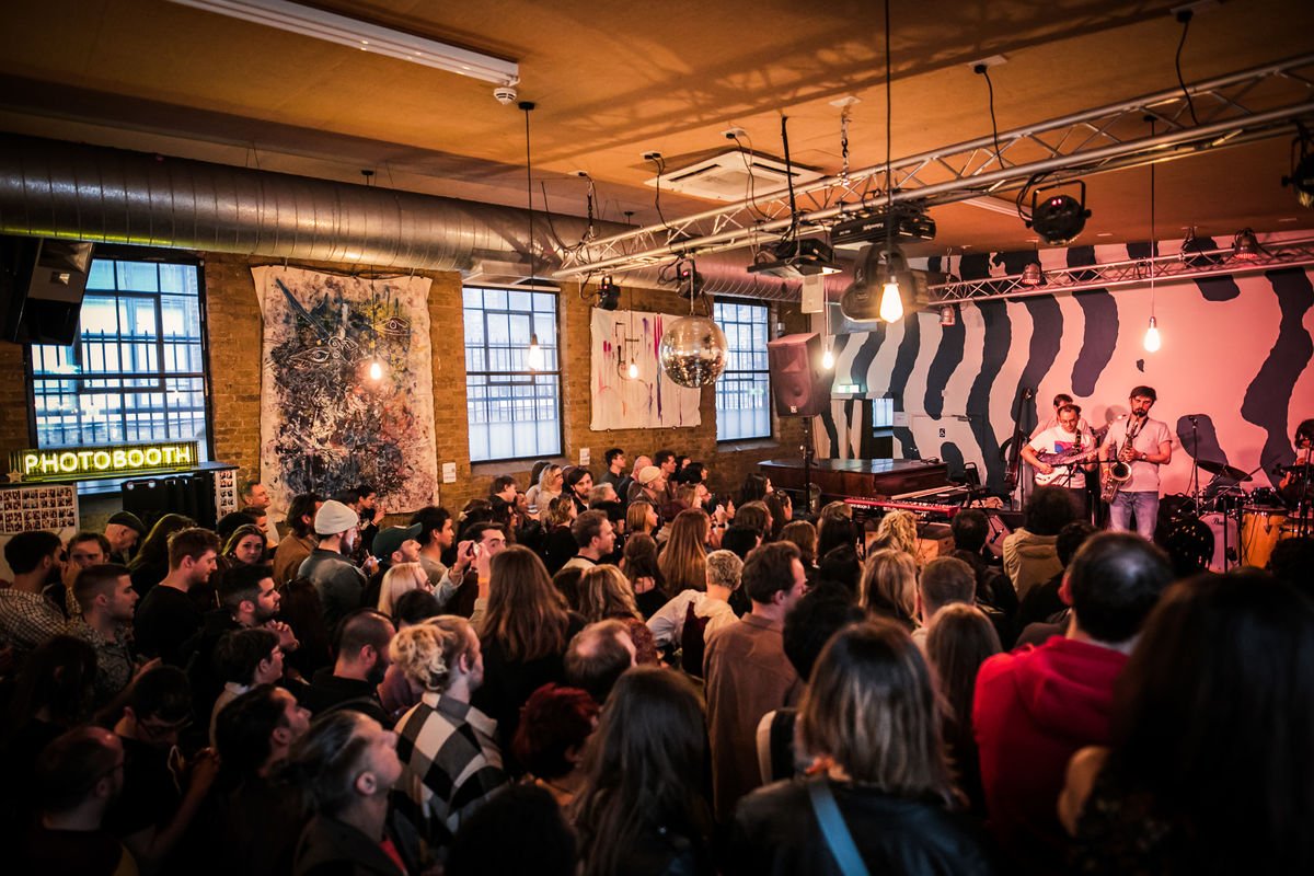 A lively concert event in a stylish indoor venue, featuring a band performing on stage with an audience closely gathered. The decor includes artistic wall murals, a disco ball, and a photobooth sign in the background.