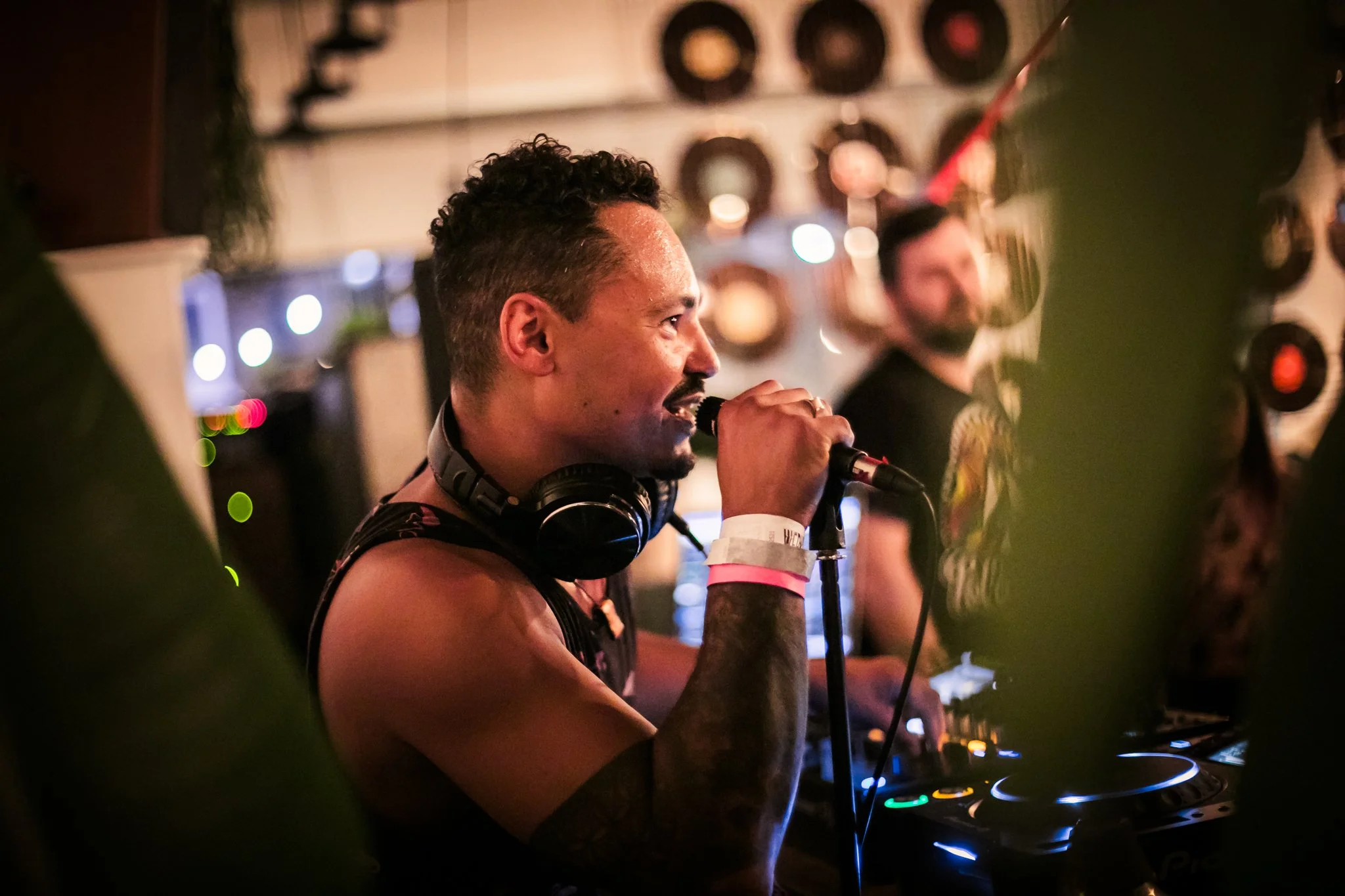 DJ performing at a lively event with headphones around neck and microphone in hand, surrounded by blurred decorative lights and records on wall.