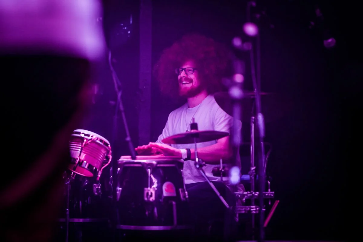 Musician playing conga drums on stage under purple lighting.