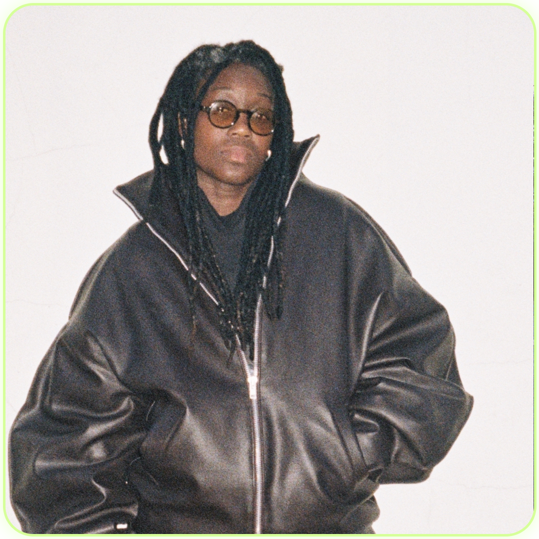 Person with dark braided hair, wearing glasses and a black leather jacket, standing in front of a plain light-colored wall.