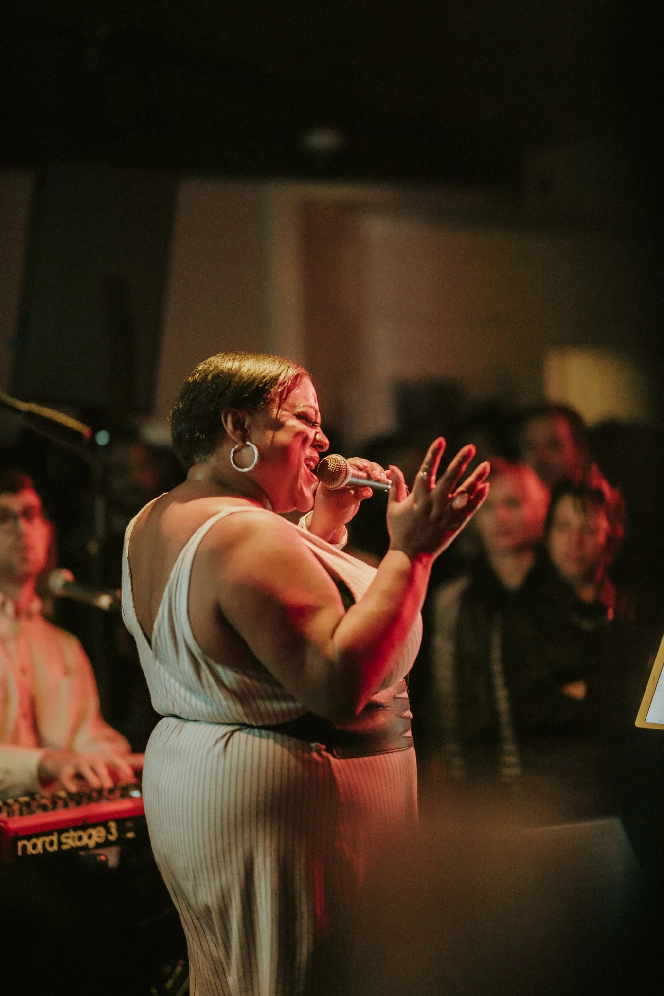 Female singer passionately performing on stage with microphone, pianist in background, and audience watching.