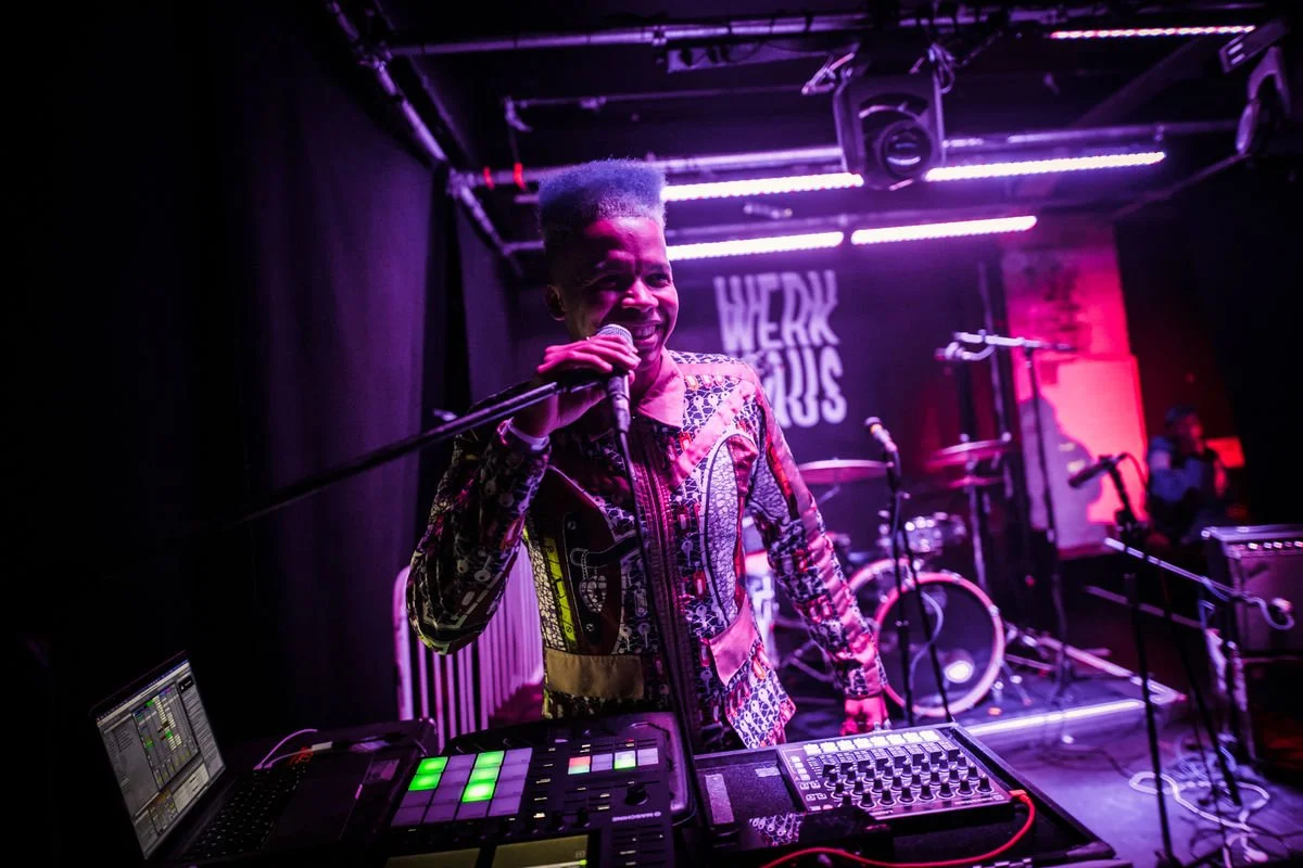 Musician performing on stage with equipment and microphone, colorful lighting, "WERK MODUS" logo in background.