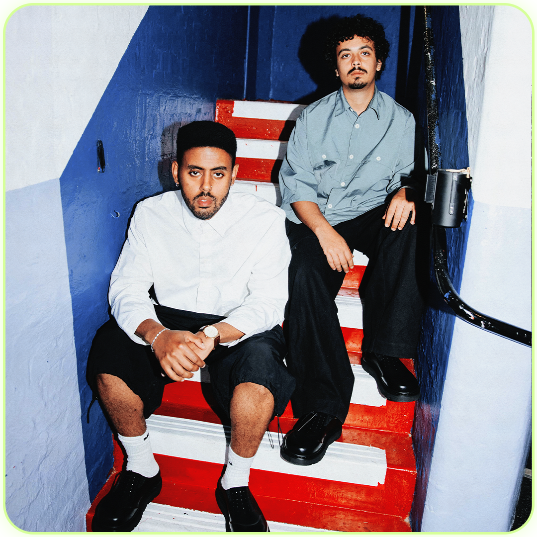 Two young men sitting on a colorful staircase with blue walls. The man on the left has dark hair, a beard, and is wearing a white shirt, black shorts, white socks, and black shoes. The man on the right has curly black hair and a mustache, wearing a light gray shirt and black pants.