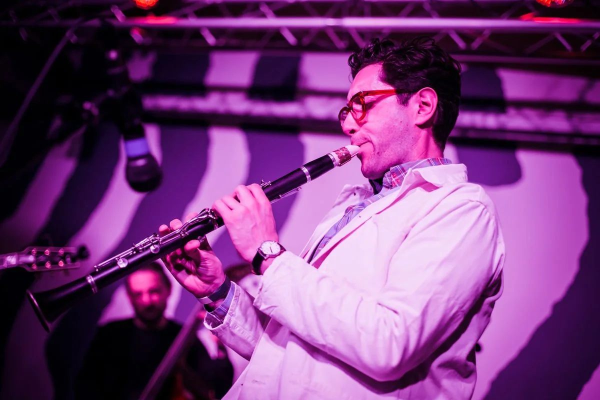 Person playing clarinet on stage with microphone overhead, pink lighting, and audience in background.