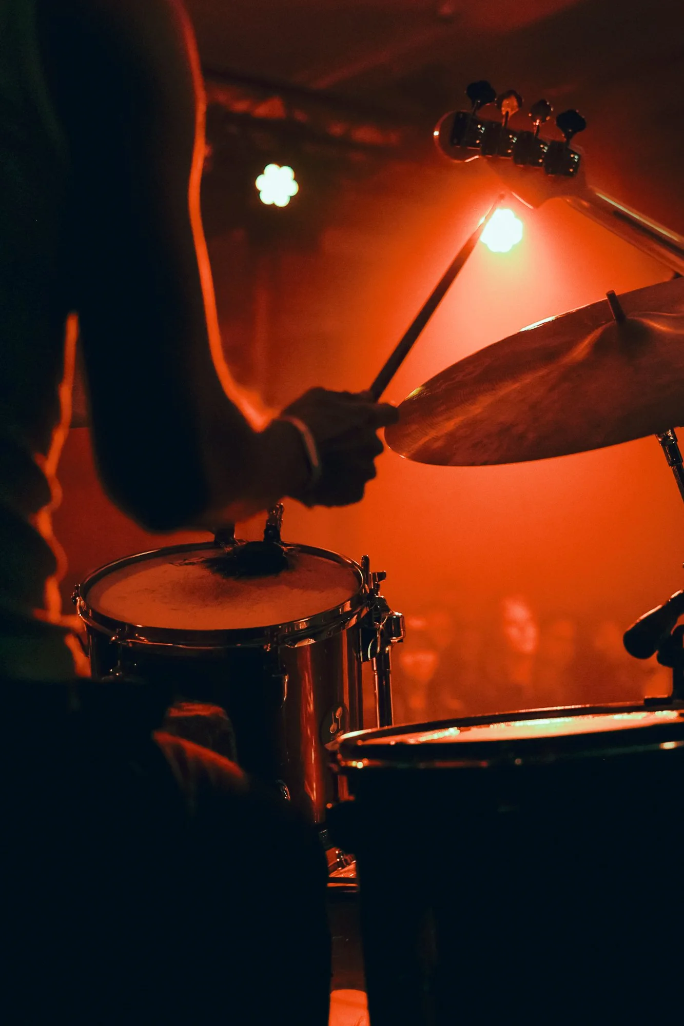 Drummer playing drums in a dimly lit concert venue with audience visible in the background