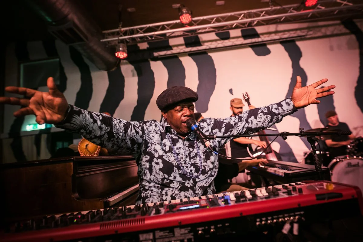 Musician playing keyboard and singing at a live jazz performance, with arms outstretched, backed by a band.