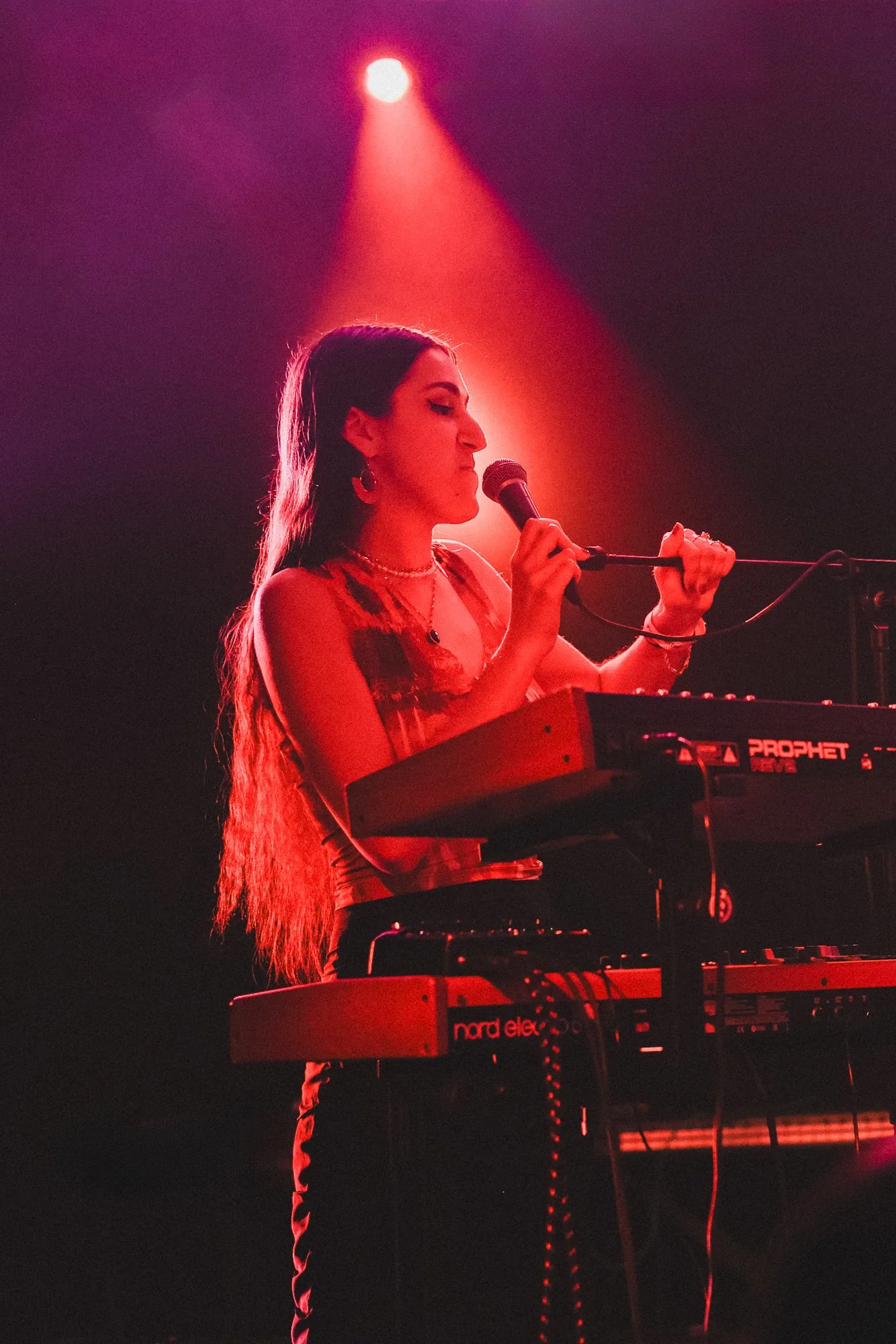 Female musician singing and playing keyboard on stage, red lighting, live performance.