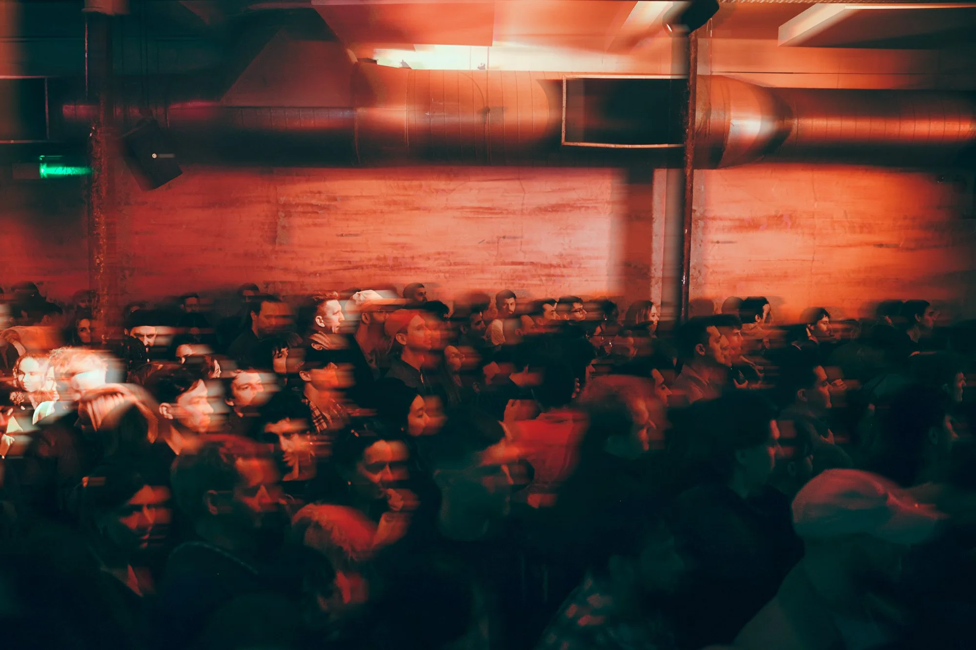 Blurred crowd in a dimly lit indoor venue with red lighting and exposed ductwork.