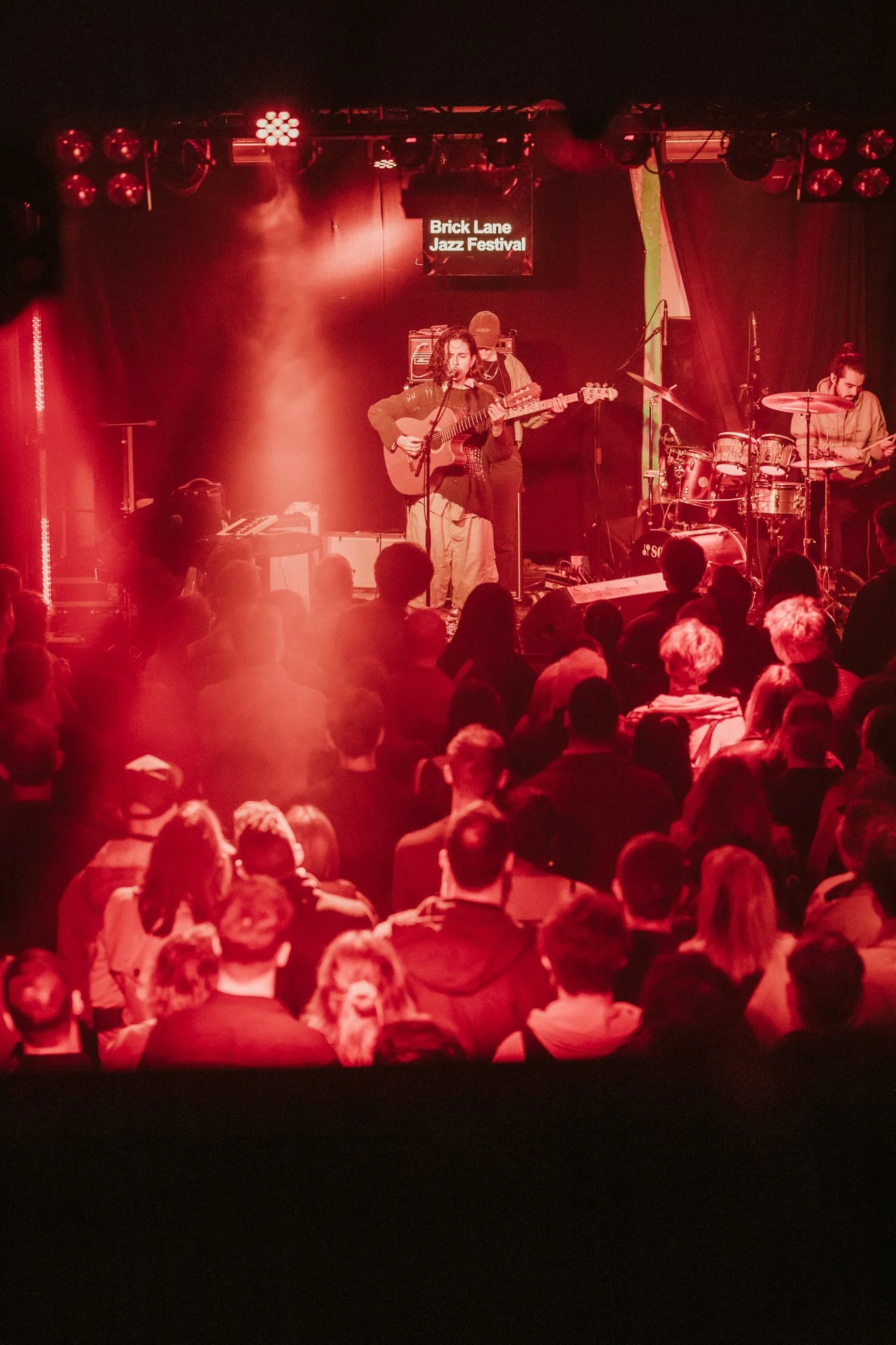 Live performance at Brick Lane Jazz Festival with musician playing guitar on stage, audience in foreground, red lighting.