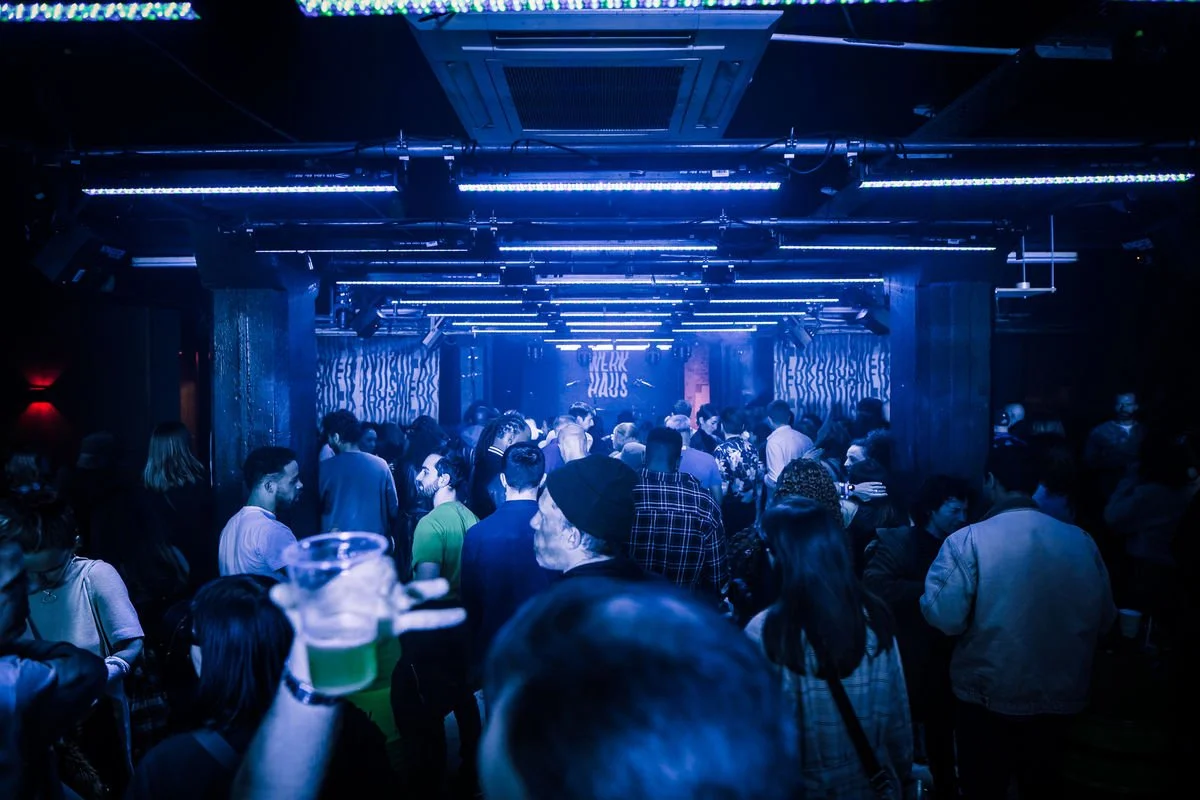Crowded nightclub interior with blue lighting, people socializing and holding drinks.