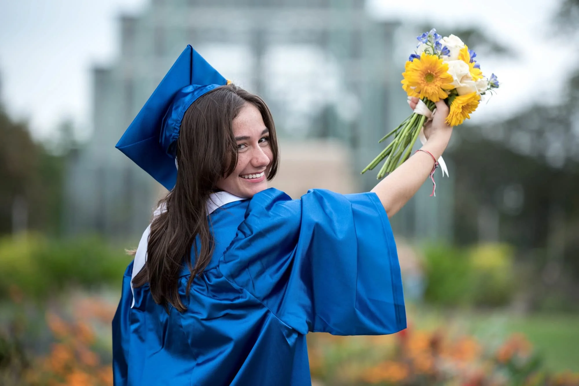 Holding a bouquet of purple, yellow, and white flowers, a high school senior girl in a blue graduation gown poses for a photo in front of the flower lined Jewel Box pathway.