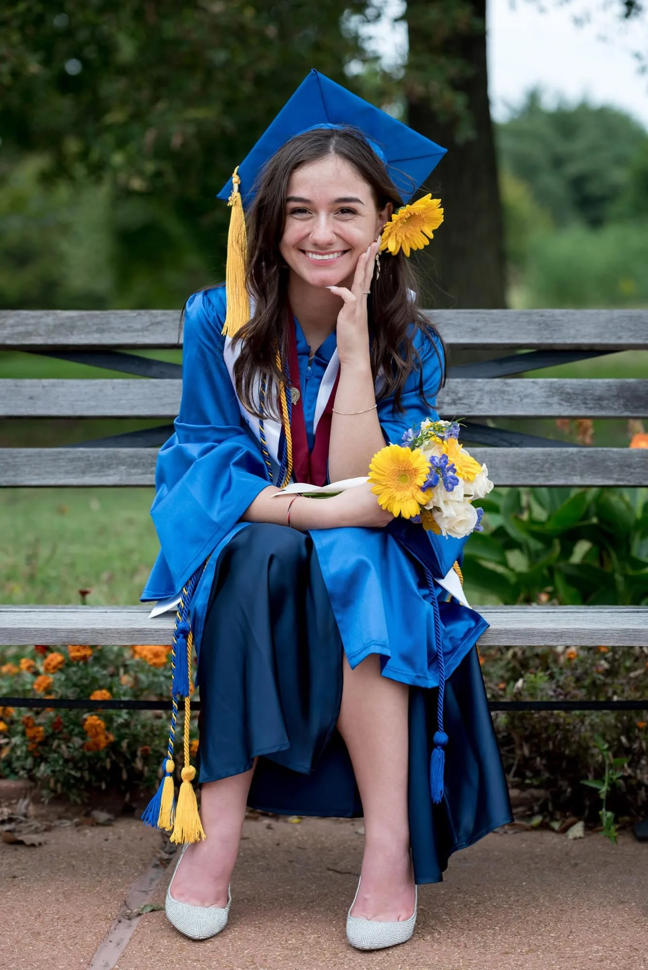 A photo shows a high school senior girl wearing a blue graduation cap and gown sitting on a wooden bench outside the Jewel Box in Forest Park.