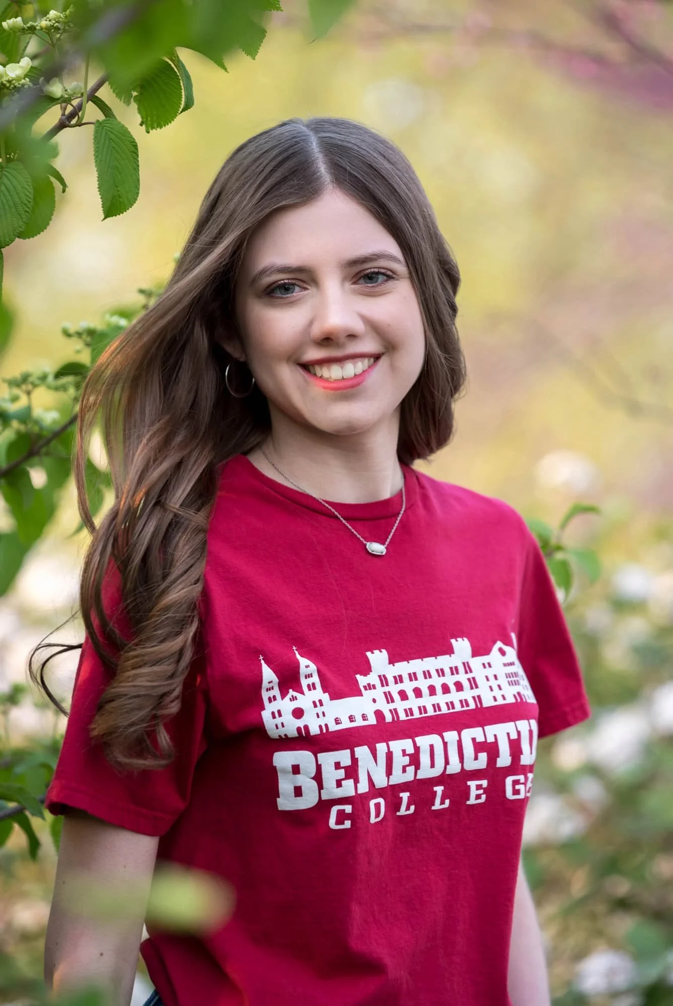 Close-up senior picture of a girl in a red college shirt with her hair blowing in the wind, posing in front of a blooming white flower bush at sunset.
