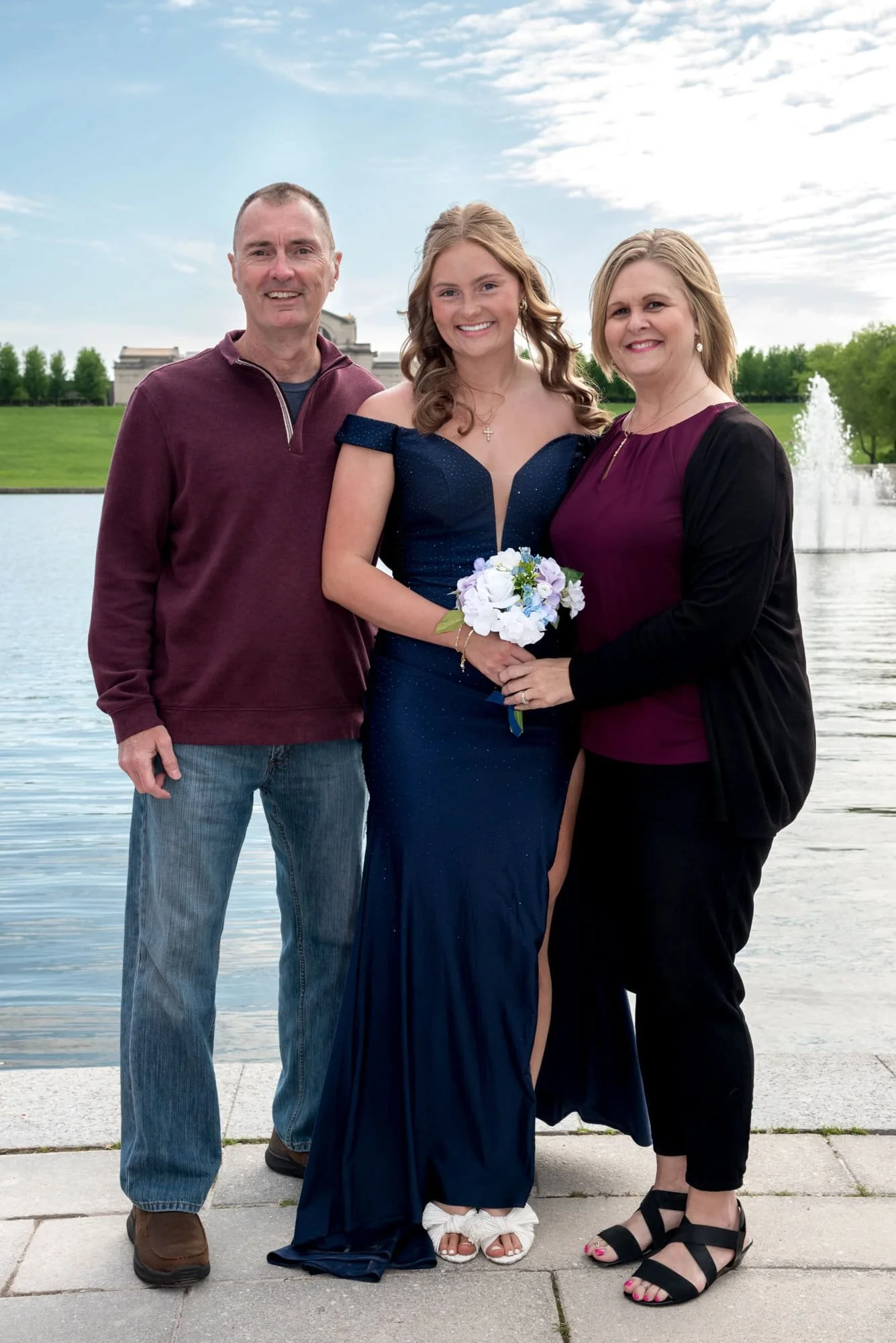 A photo captures a senior girl wearing a navy blue prom dress standing outdoors between her mom and dad at Forest Park.