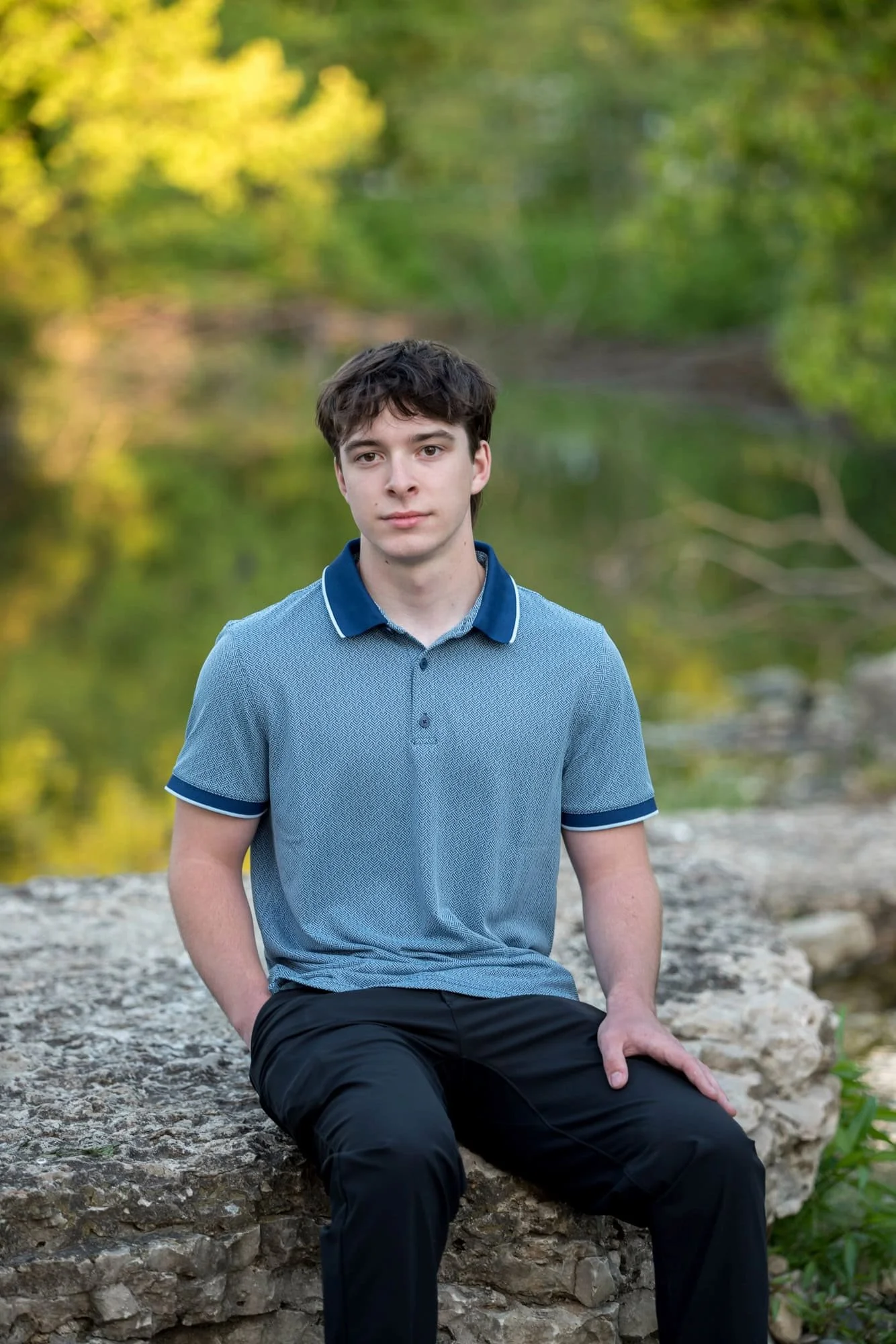 High school senior boy in a blue short-sleeve polo shirt sitting on limestone rocks along a creek bed in St. Louis.