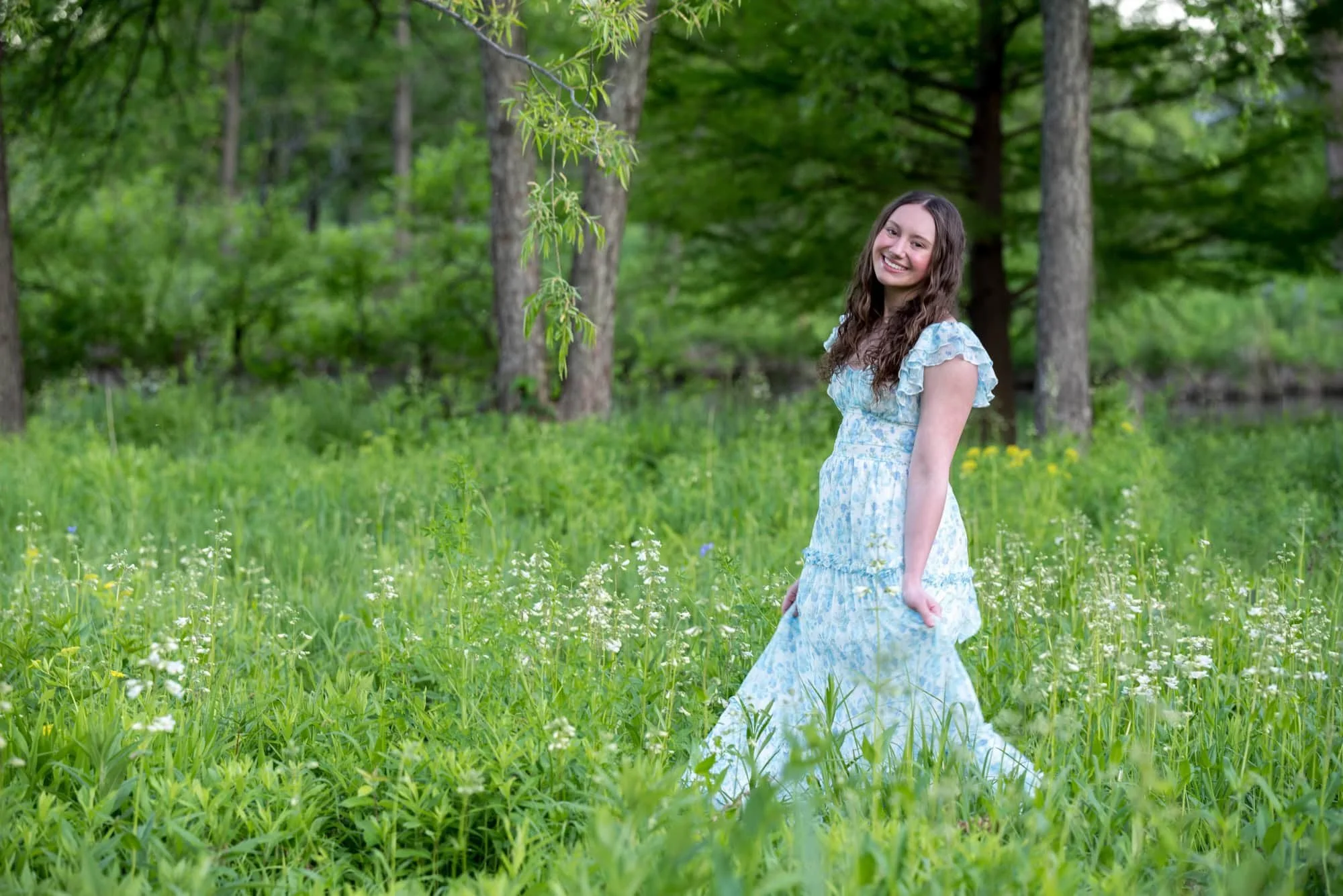 Surrounded by a lush green field of country wildflowers, a smiling senior girl walks near a wooded pond area down from Art Hill.