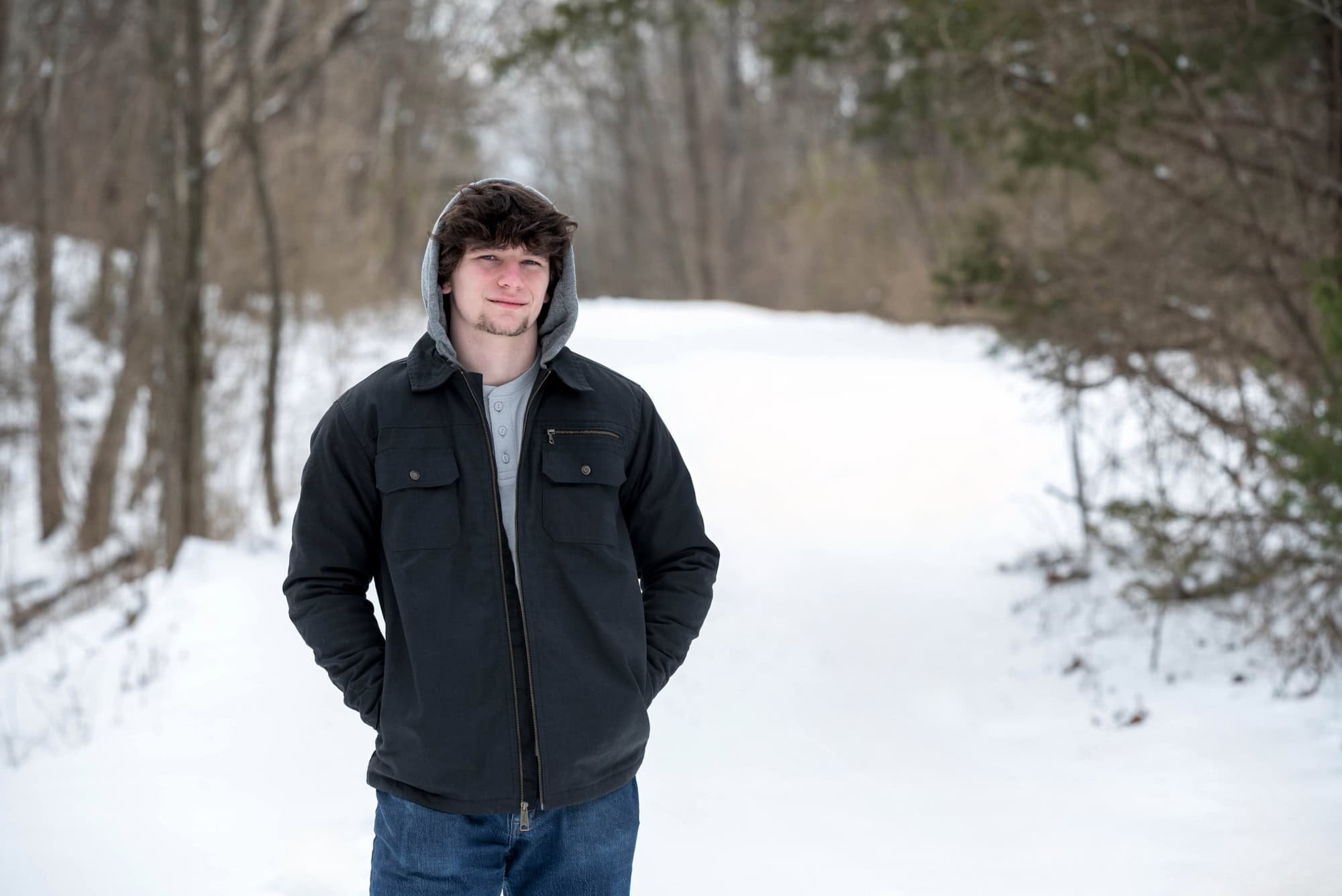 High school senior boy wearing a gray hooded sweatshirt under a black winter coat on a snowy trail in Augusta, Missouri.