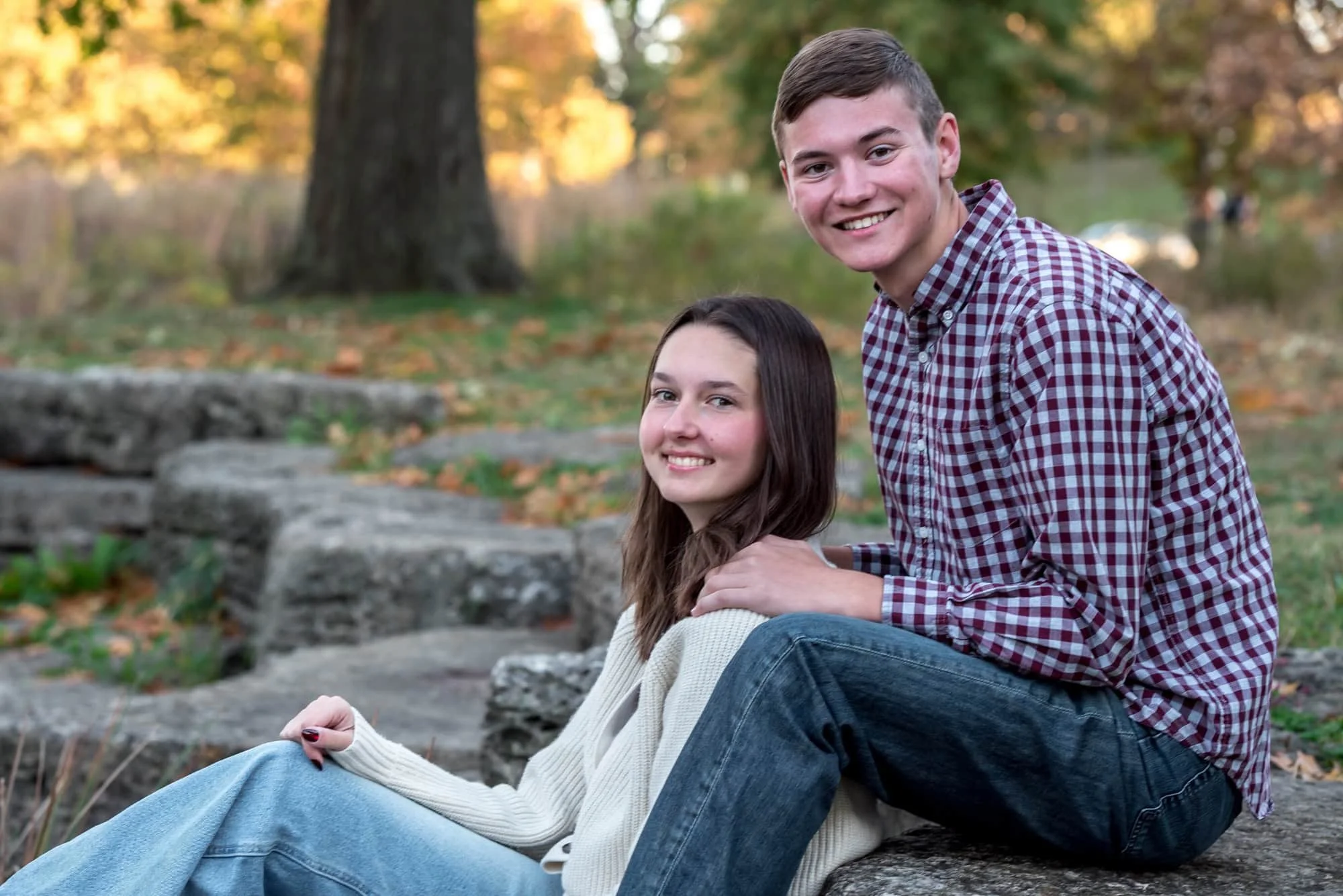 During a St. Louis fall session by the creek, a senior girl in a white sweater and her boyfriend in a maroon plaid shirt sit together on the rocks for a country style couple's photo.