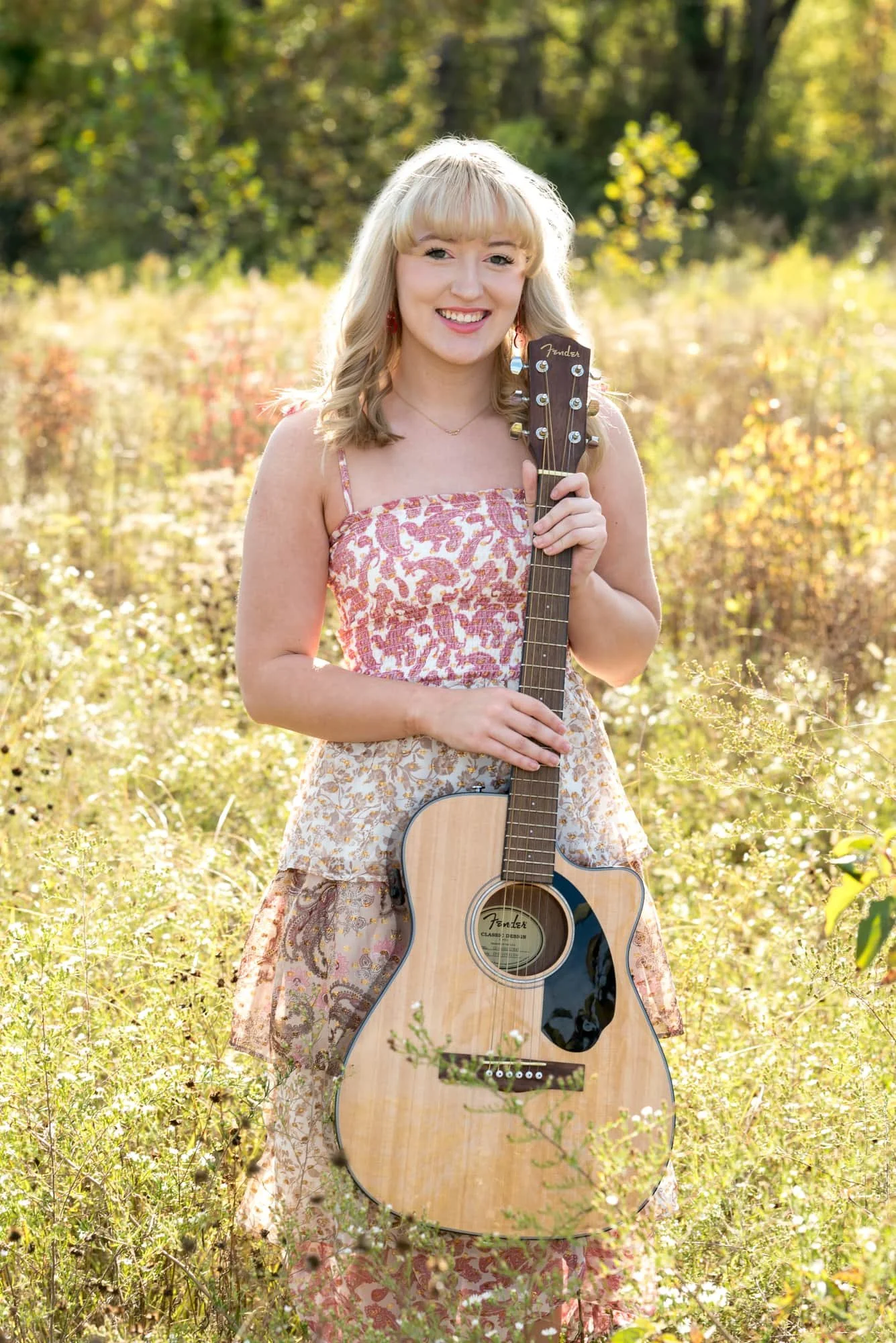 In a wildflower field at Admiral Trost Park in Columbia, Illinois, a high school senior girl wearing a pink and white floral dress holds a guitar for a fall photo.