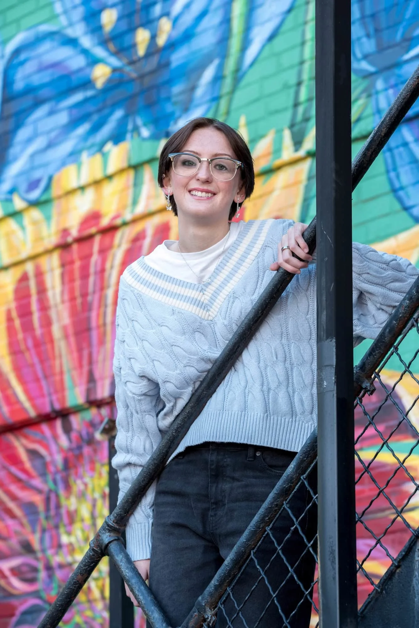 Artistic urban senior picture showing a girl in a blue sweater and black pants standing on a black fire escape stairway with a colorful flower mural in the background.