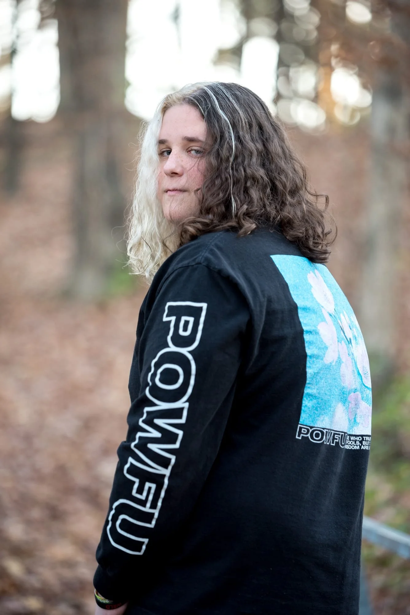 Serious close-up senior portrait of a boy looking back over his shoulder in the woods, showcasing his unique split-colored hair and the band logo on his black sweatshirt in Waterloo, IL.