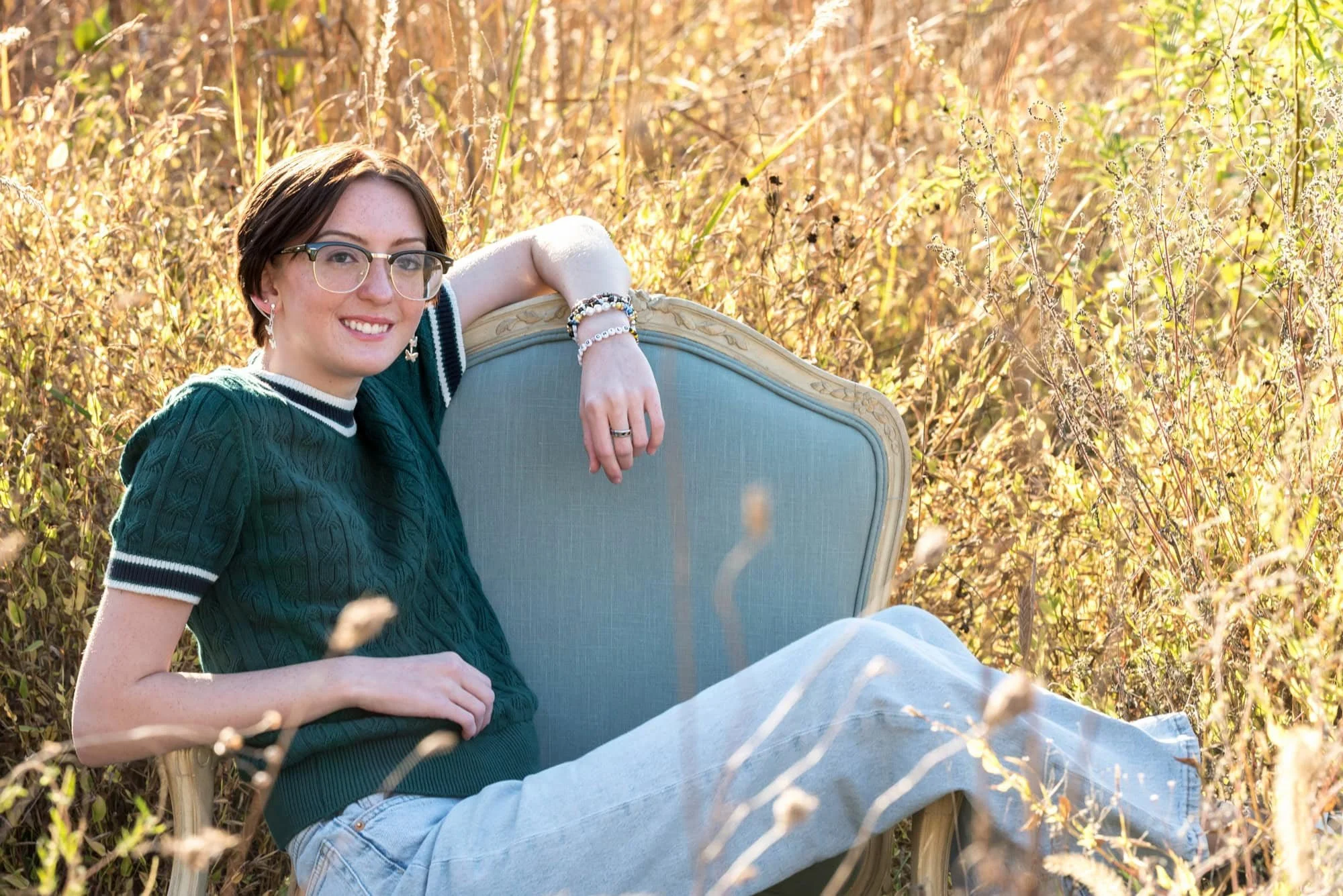 Relaxed park senior picture of a girl wearing a green short-sleeve sweatshirt and blue jeans, lounging in a blue chair with her feet kicked over the armrest at Queeny Park.