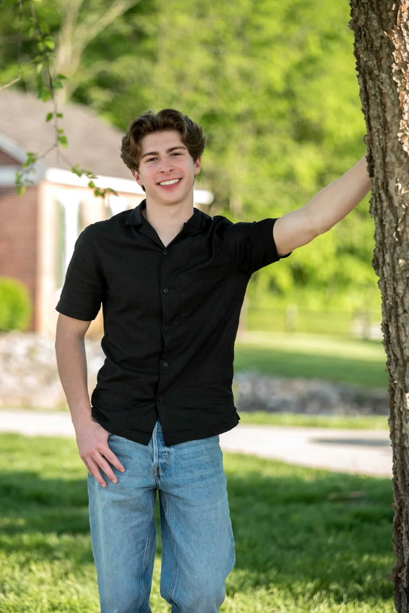 A picture captures a high school senior guy wearing a black shirt and blue jeans leaning against a tree in Columbia, Illinois.