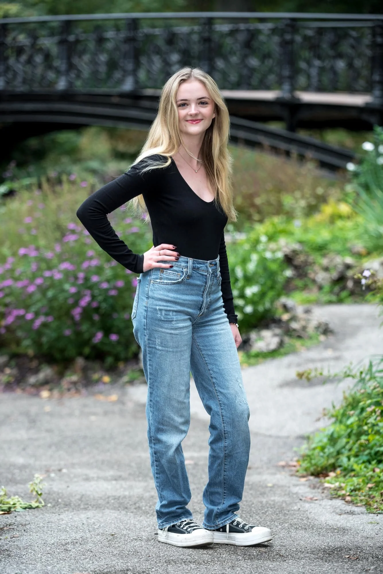 Whimsical senior photo of a girl wearing a black long-sleeve shirt, blue jeans, and black Converse sneakers, posing with her hand on her hip on a pathway framed by a historic black bridge and purple spring flowers at Lafayette Park.