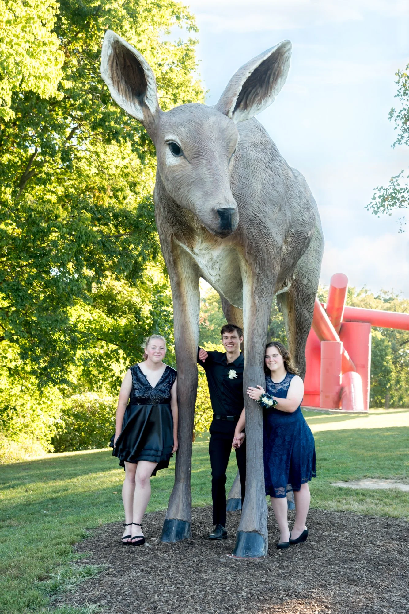 Bright, sunlit group photo of three friends standing beneath the massive deer sculpture with glowing green trees and a geometric red sculpture framing the background taken by Sunset Hills photographer.