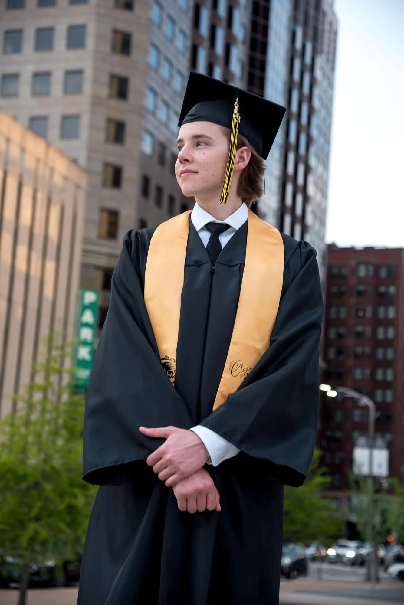 High school senior boy in his graduation cap and gown, looking off into the distance on a downtown St. Louis street corner with tall city buildings blurred in the background.