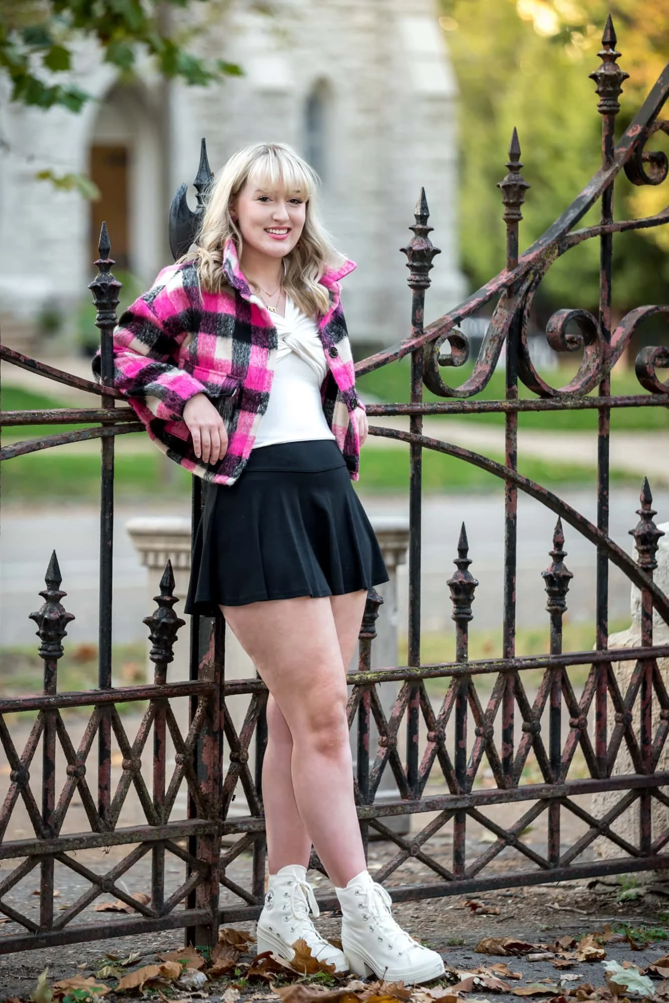 A senior girl leans back against a rod iron fence in Lafayette Park in front of a historic building wearing a pink plaid jacket, white shirt, black shirt, and blue shoes for a photo.