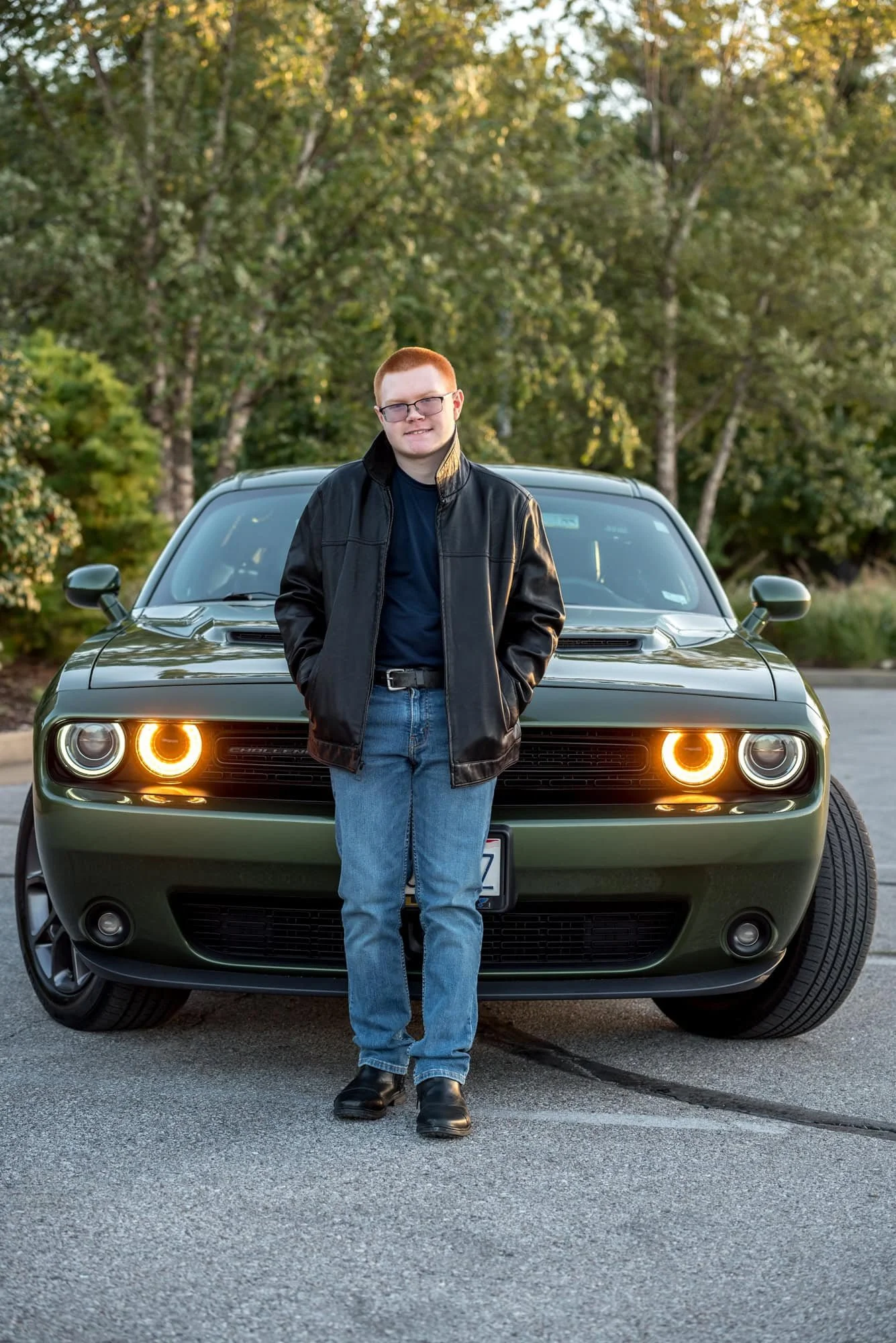 Sharp automotive senior picture showing a boy in a black leather jacket standing against the hood of his green Dodge Challenger in Forest Park.