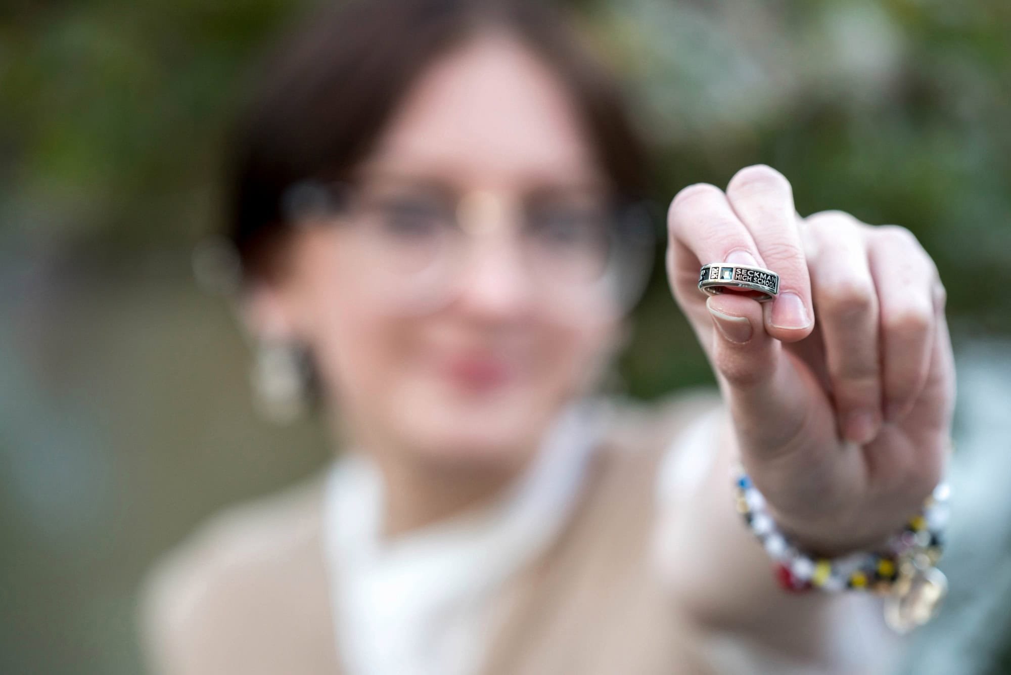 Creative close-up senior photo focusing on a girl's hand holding out her Seckman High School class ring, with her smiling face softly blurred in the background at Queeny Park.