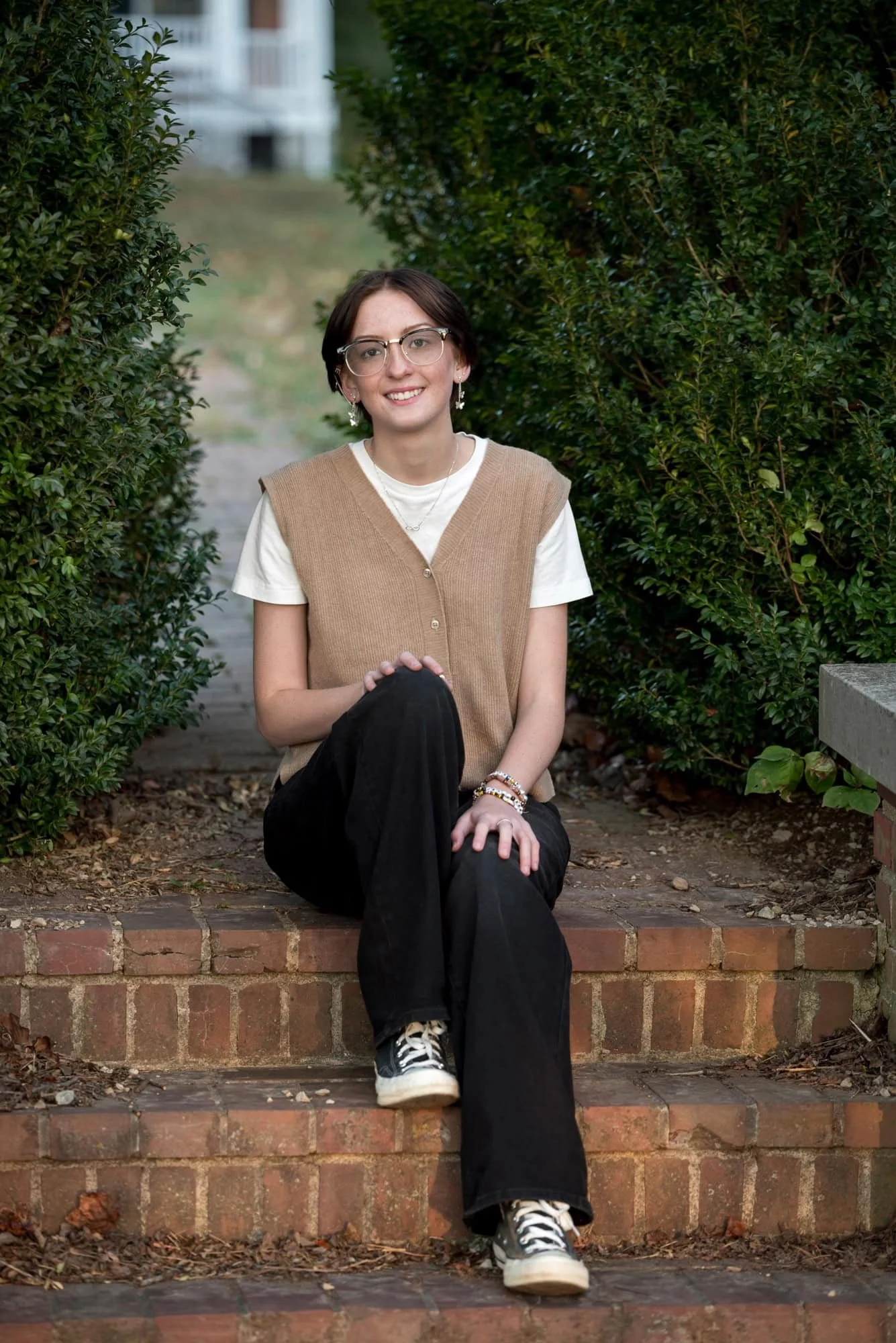 Casual outdoor senior photo taken by a St. Louis photographer, showing a girl in a tan vest, white t-shirt, black pants, and black Converse sitting on brick steps with her hands on her knees.