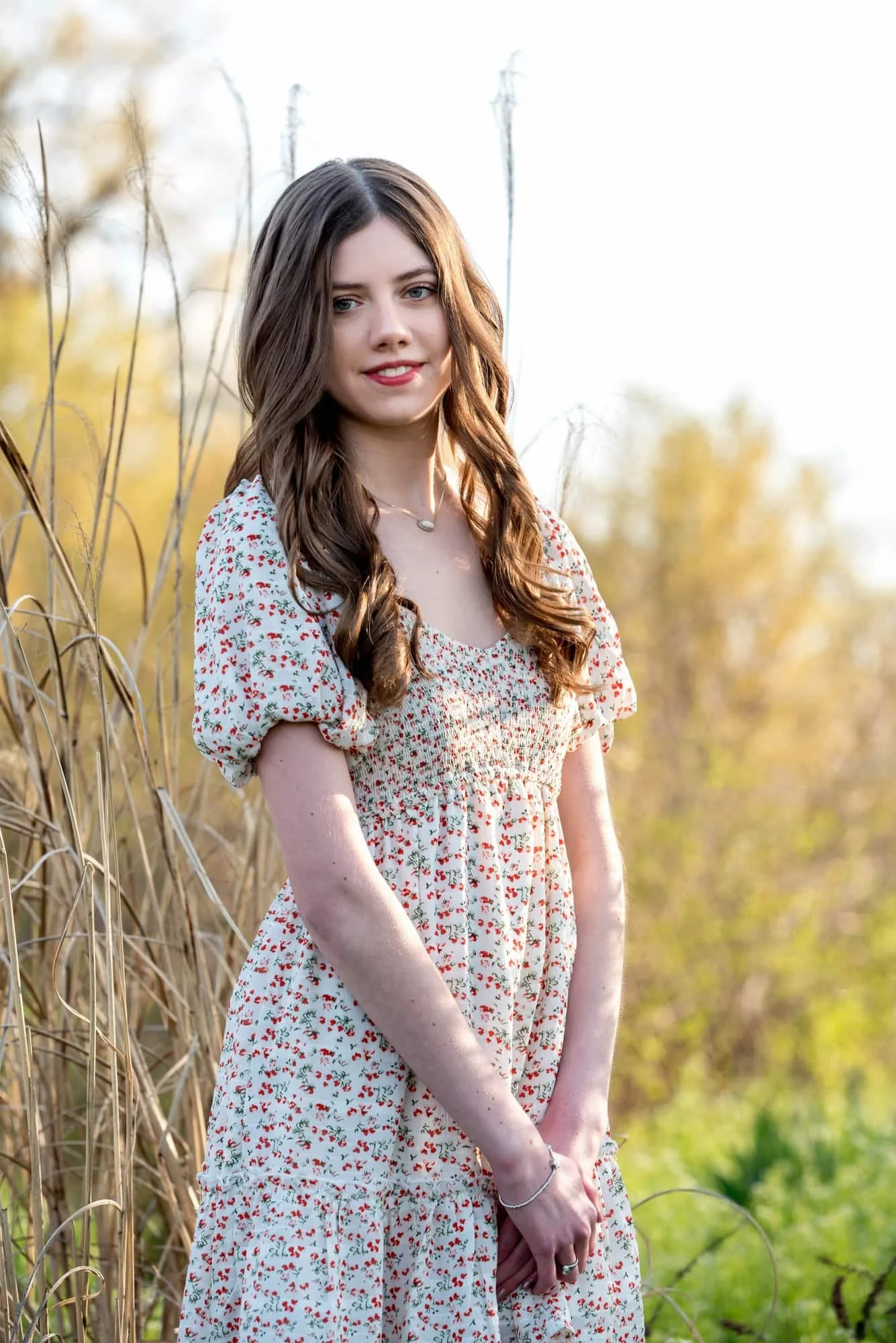 Senior girl wearing a white dress with red flowers, standing in front of tall rustic grasses lit up by a soft, warm spring sunset at Forest Park.