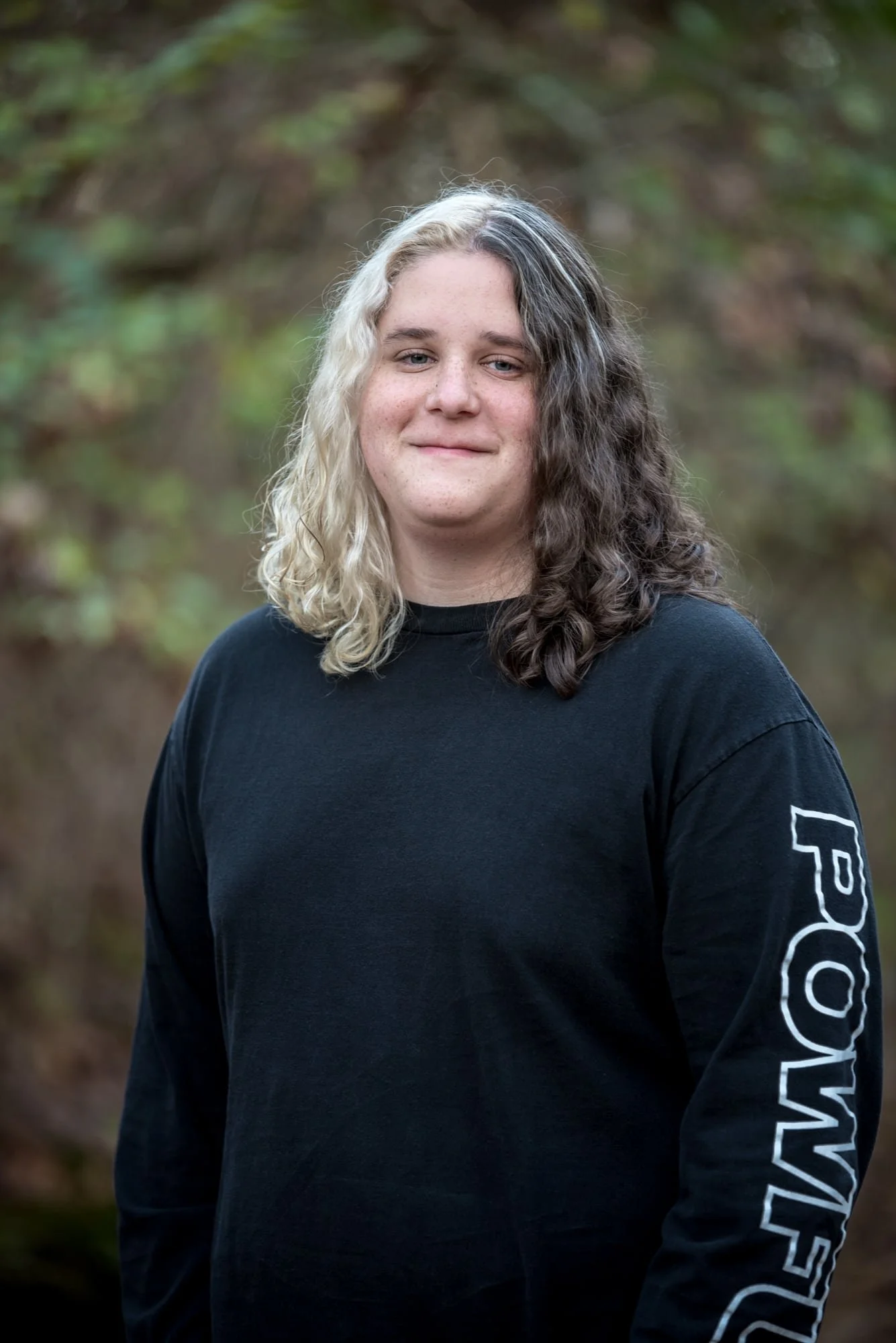 Senior guy wearing a casual black sweatshirt, smiling in front of a lush green foliage backdrop during a fall high school senior photoshoot at Konarcik Park.