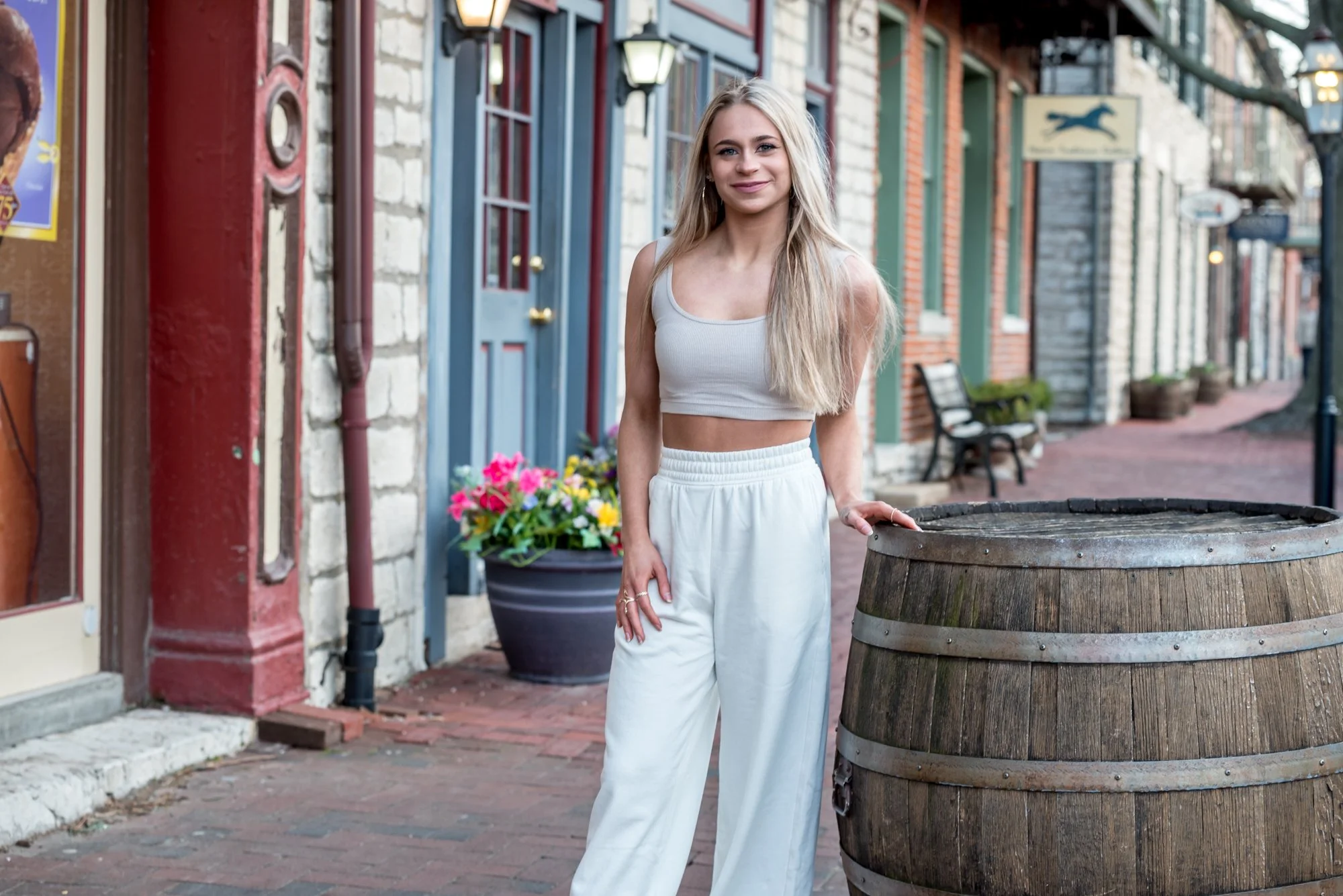 High school senior girl wearing a casual tan crop top and white sweatpants, standing with her hand on a rustic barrel along the historic brick walkway lined with colorful storefronts in St. Charles MO.
