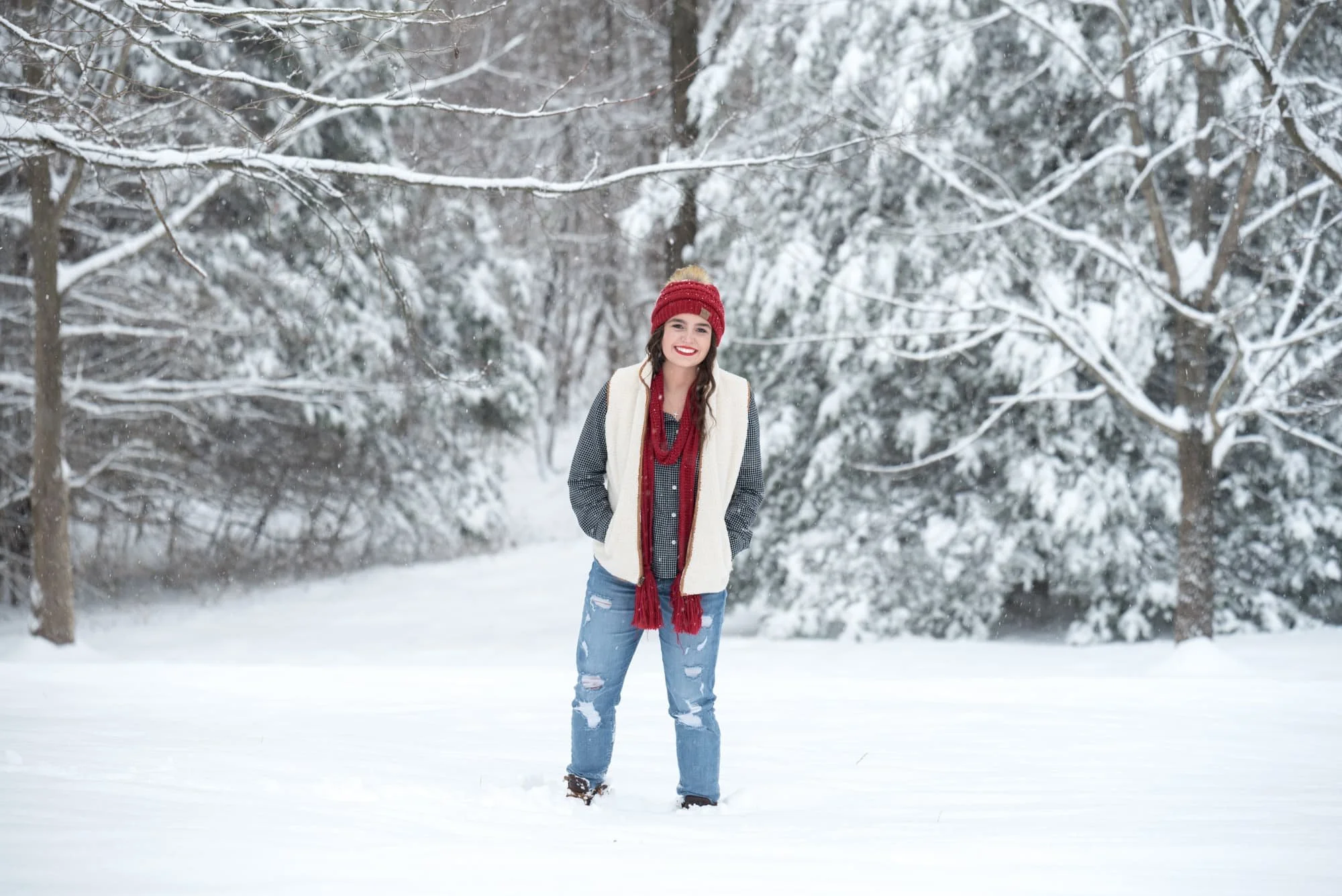 High school senior girl standing with her hands in her pockets in the middle of a wide snowy field, wearing a red beanie, white vest, and red scarf with snow-covered trees behind her in Columbia IL.