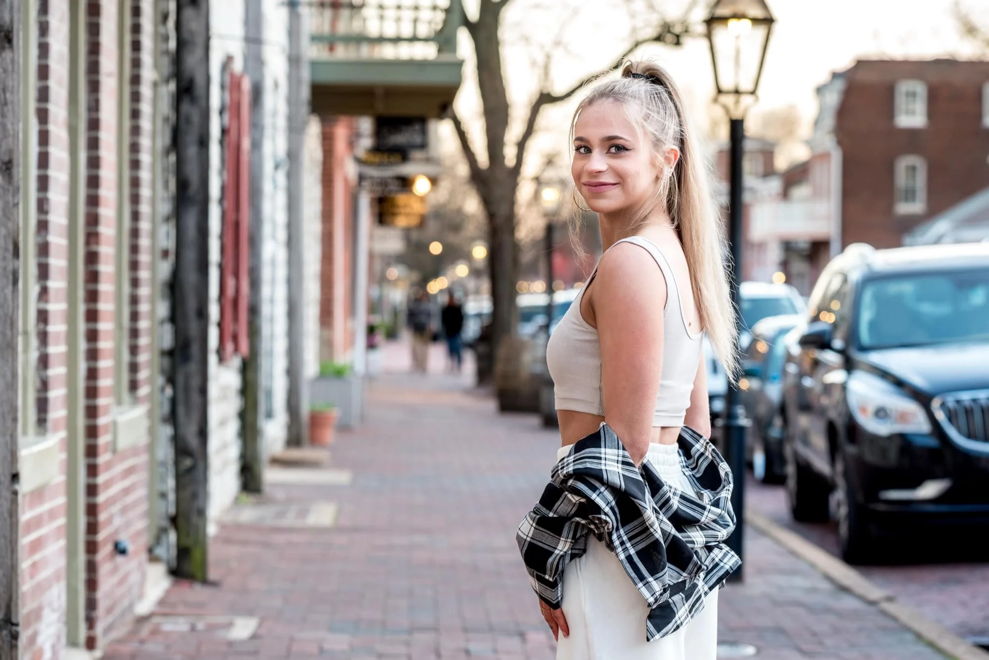 A high school senior girl with a ponytail wearing a cream crop top and white pants standing on a brick sidewalk during a senior portrait session on Main Street St. Charles.