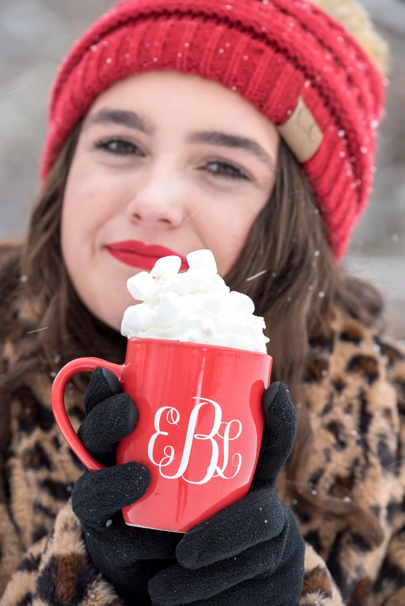 Close-up senior photo of a girl in a leopard coat, black gloves, and a red beanie, holding out an overfilled red monogrammed mug of hot chocolate and giant marshmallows as snow falls around her.