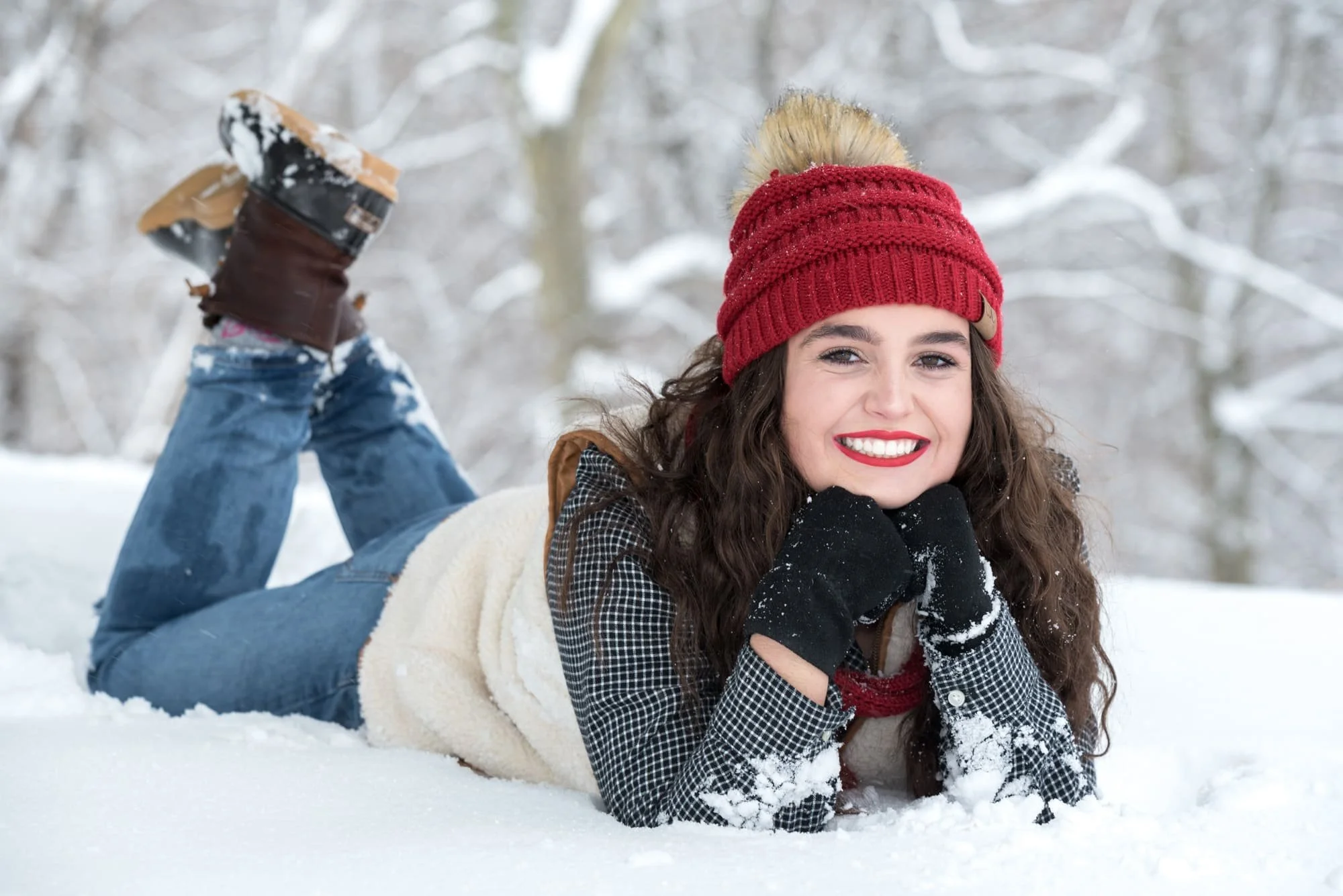 Senior picture of a girl laying in the snow with her chin resting on her hands and her feet kicked up, wearing a white vest, ripped jeans, and brown boots in Columbia IL.