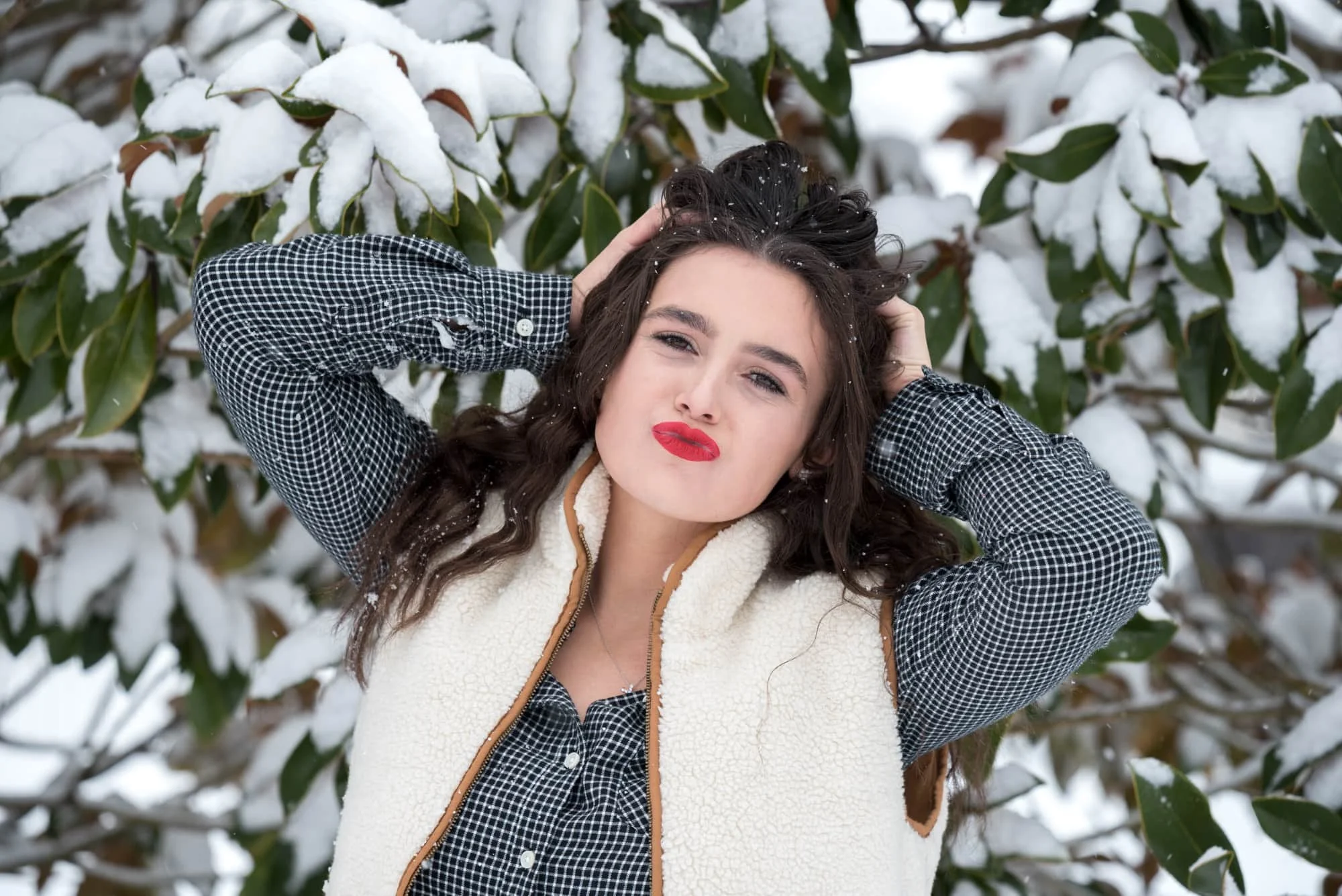 Close-up senior portrait of a girl with her hands in her hair making a playful kissy face, wearing a red beanie and white vest in front of snow-covered green leaves.